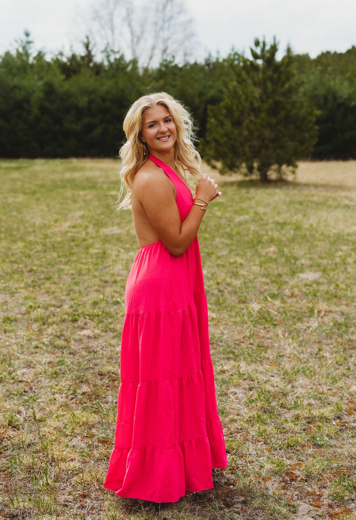 Woman in hot pink halter maxi dress posing outdoors in a grassy field with evergreen trees in background.