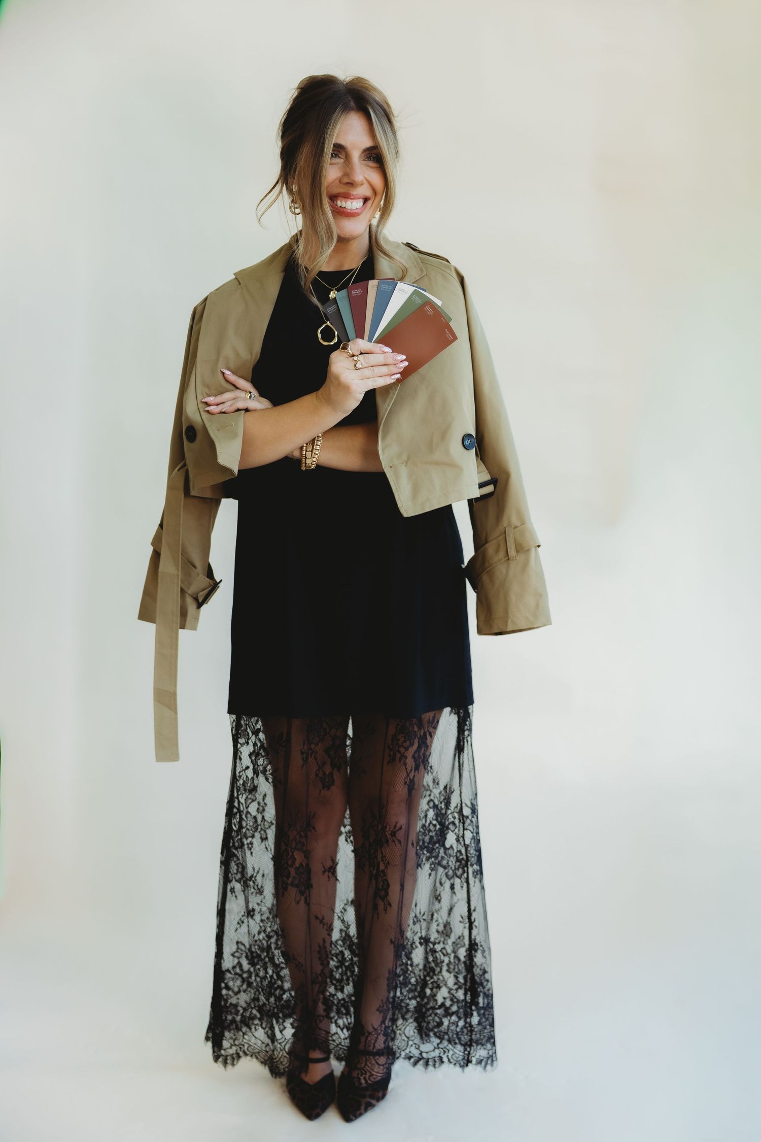 Smiling woman in a tan trench jacket and black lace maxi skirt holding a fan of color swatches against a plain backdrop.