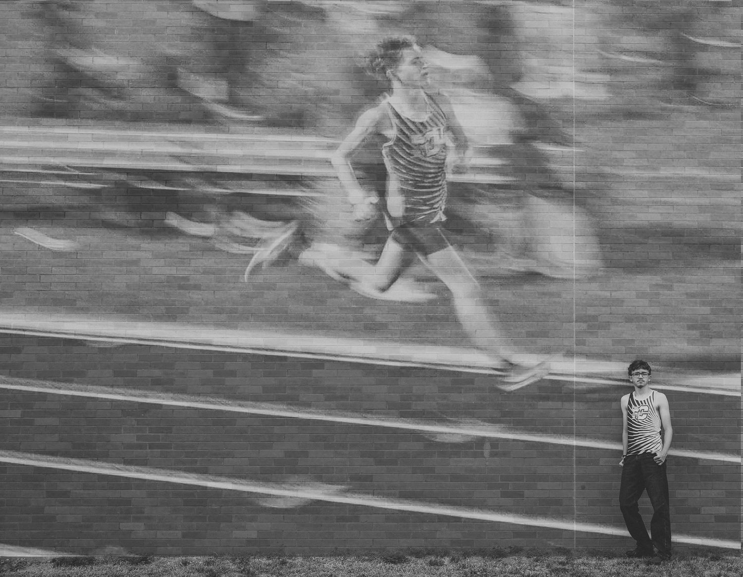 Motion-blurred runner sprints along a track while a shirtless spectator stands calmly in the foreground in this black and white photo.
