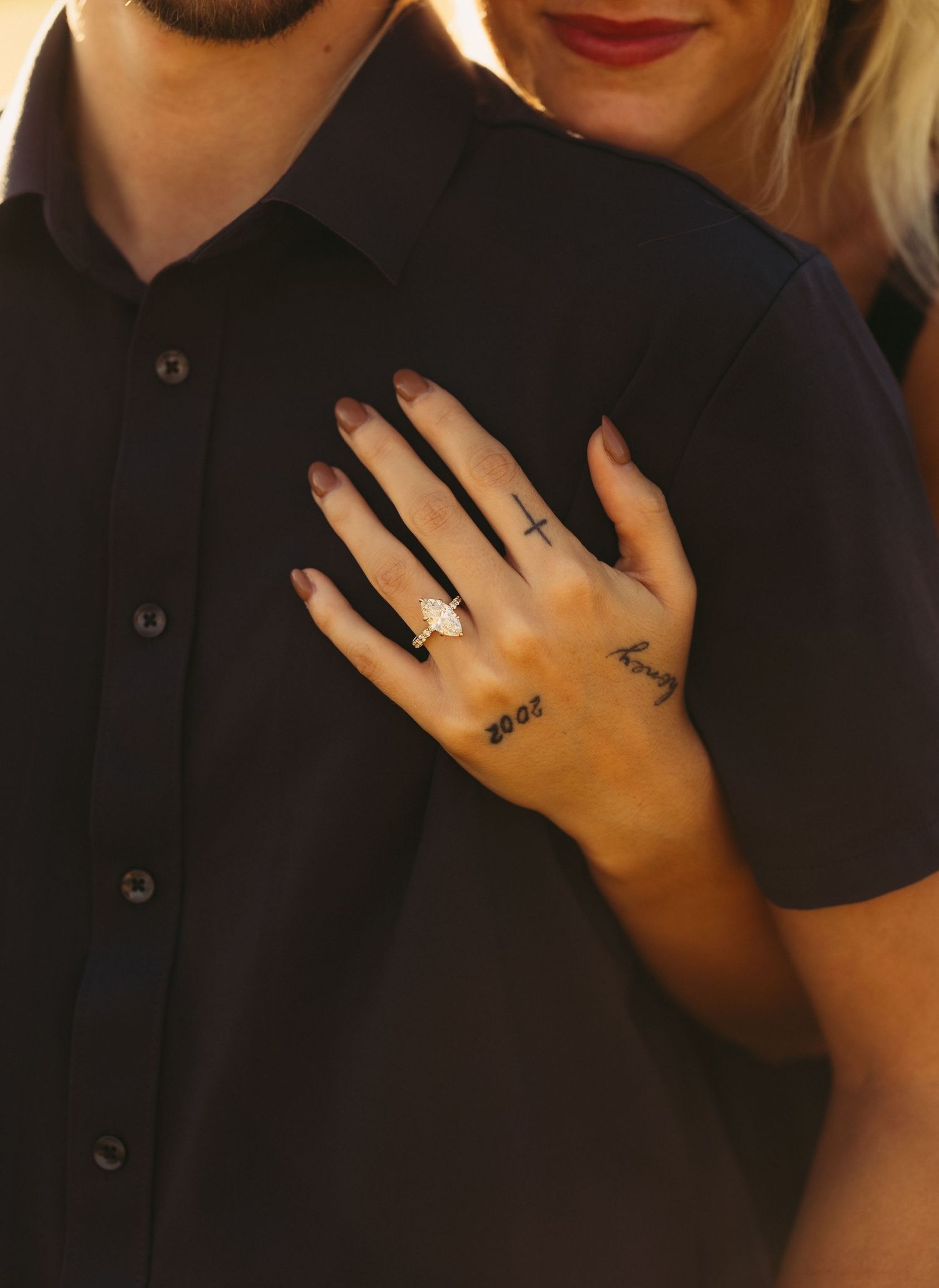 Woman's hand with small tattoos and an oval diamond engagement ring resting on a man's dark button-up shirt during a warm sunset engagement.