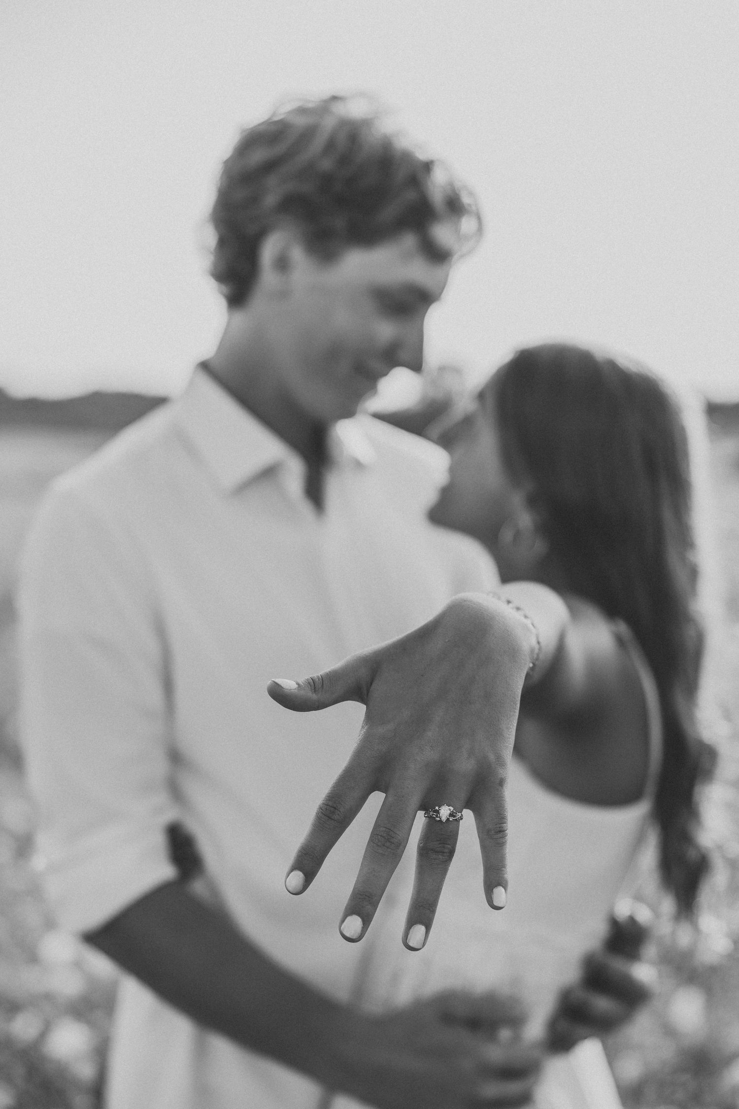 Black and white photo of couple nearly kissing while showing off engagement ring toward camera.