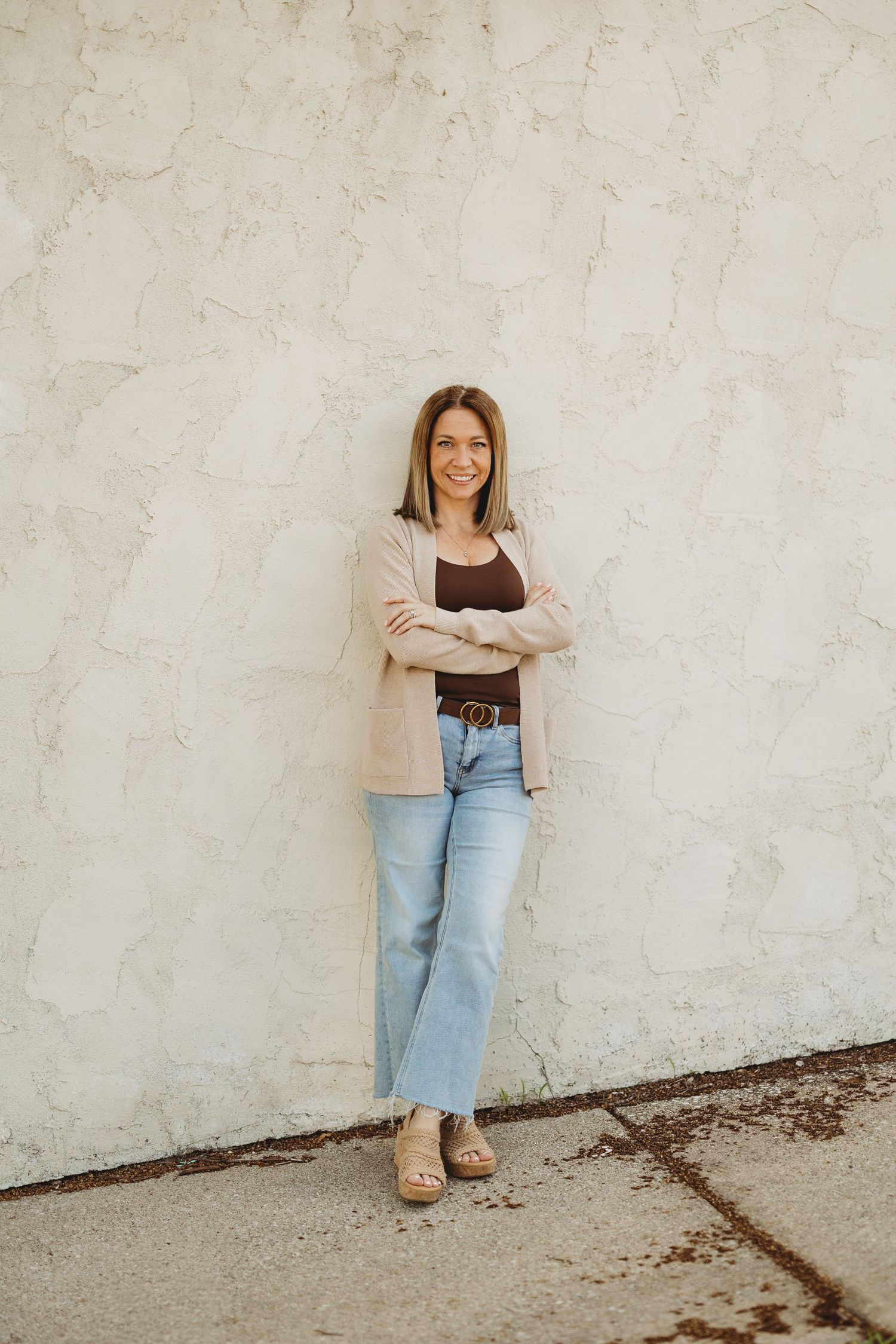 Woman in light blazer and jeans smiling with arms crossed against a white textured wall.