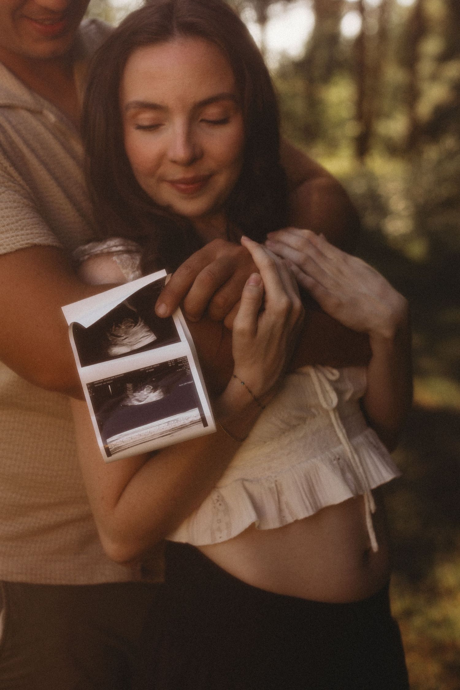 Pregnant woman and partner holding ultrasound photos together in warm outdoor setting.