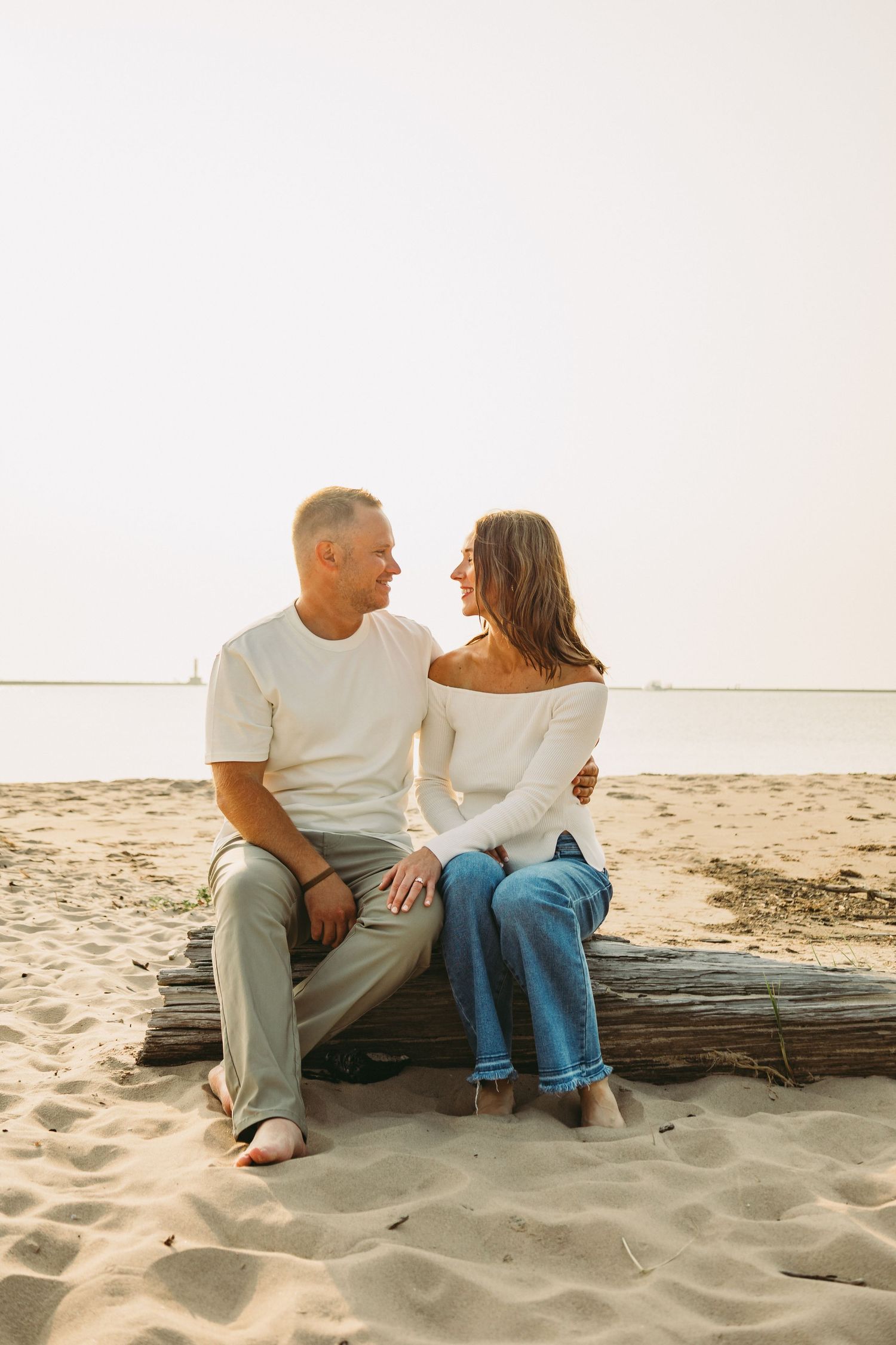 A couple sits closely on driftwood at a sandy beach, smiling at each other in casual white tops and jeans.