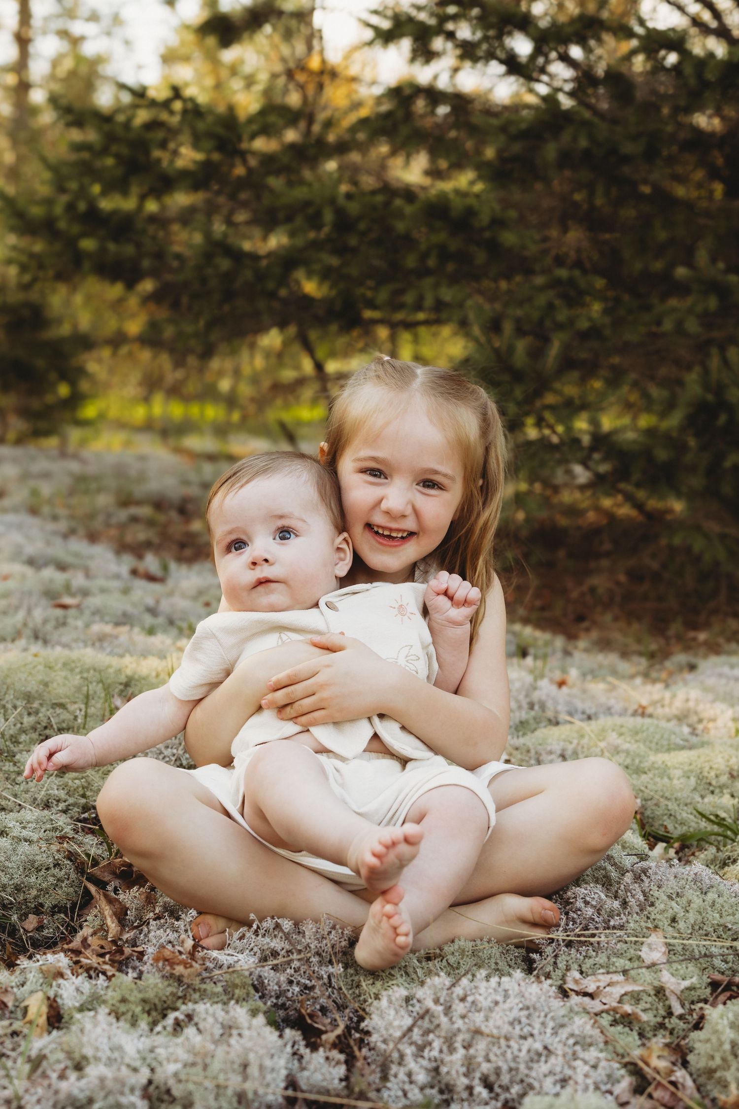 Young girl smiling and hugging baby sibling outdoors on mossy ground with evergreen trees in background.