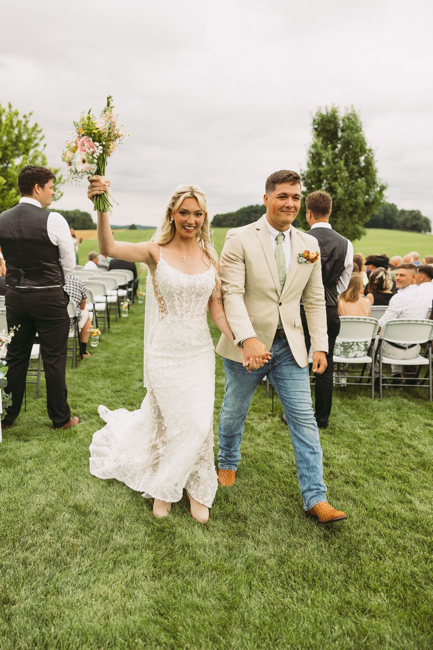 Newlyweds celebrate while walking down the aisle at an outdoor wedding ceremony with guests seated on white chairs.