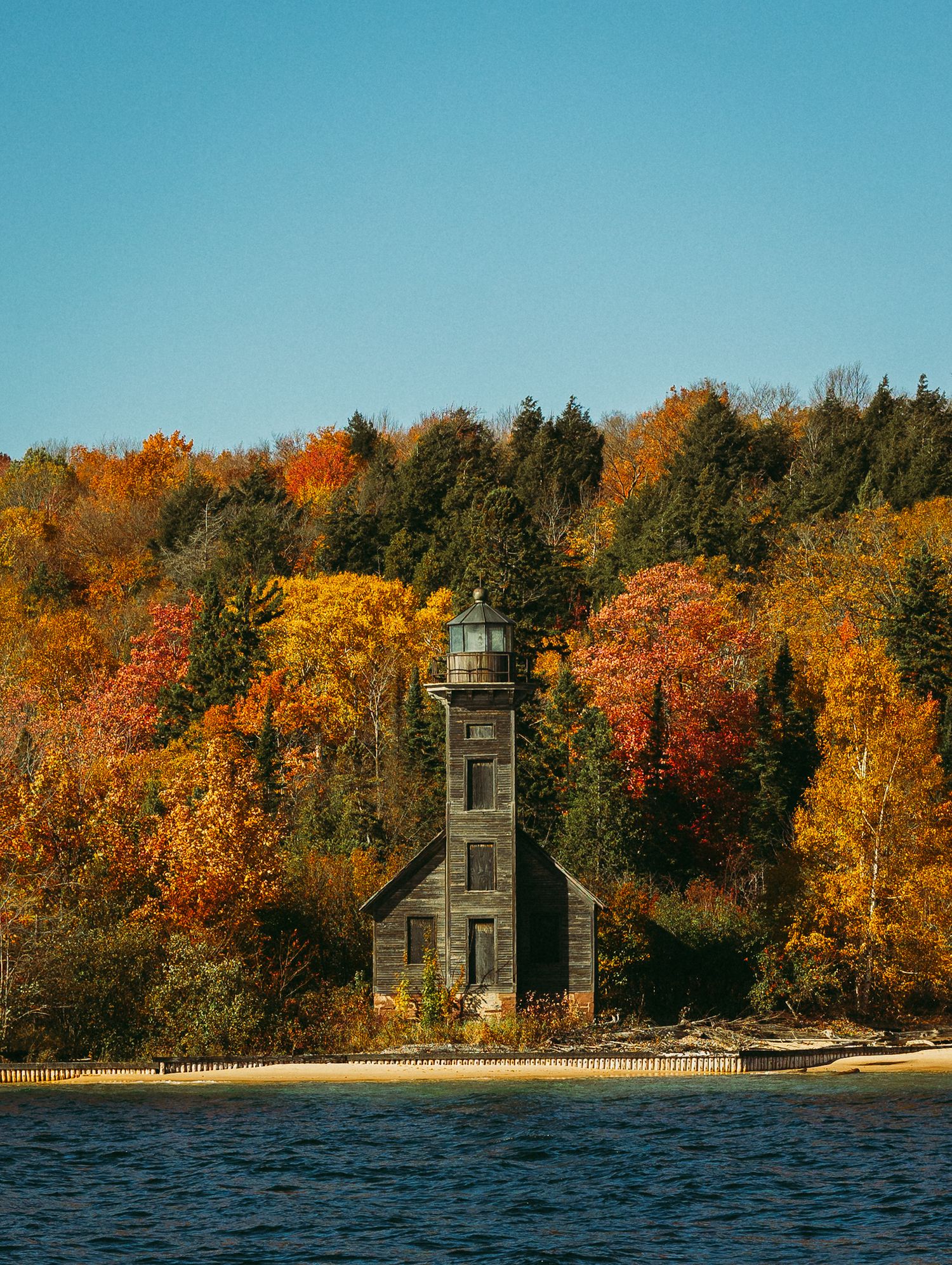Autumn trees with vibrant orange foliage surround a wooden lighthouse structure on a lake shore.