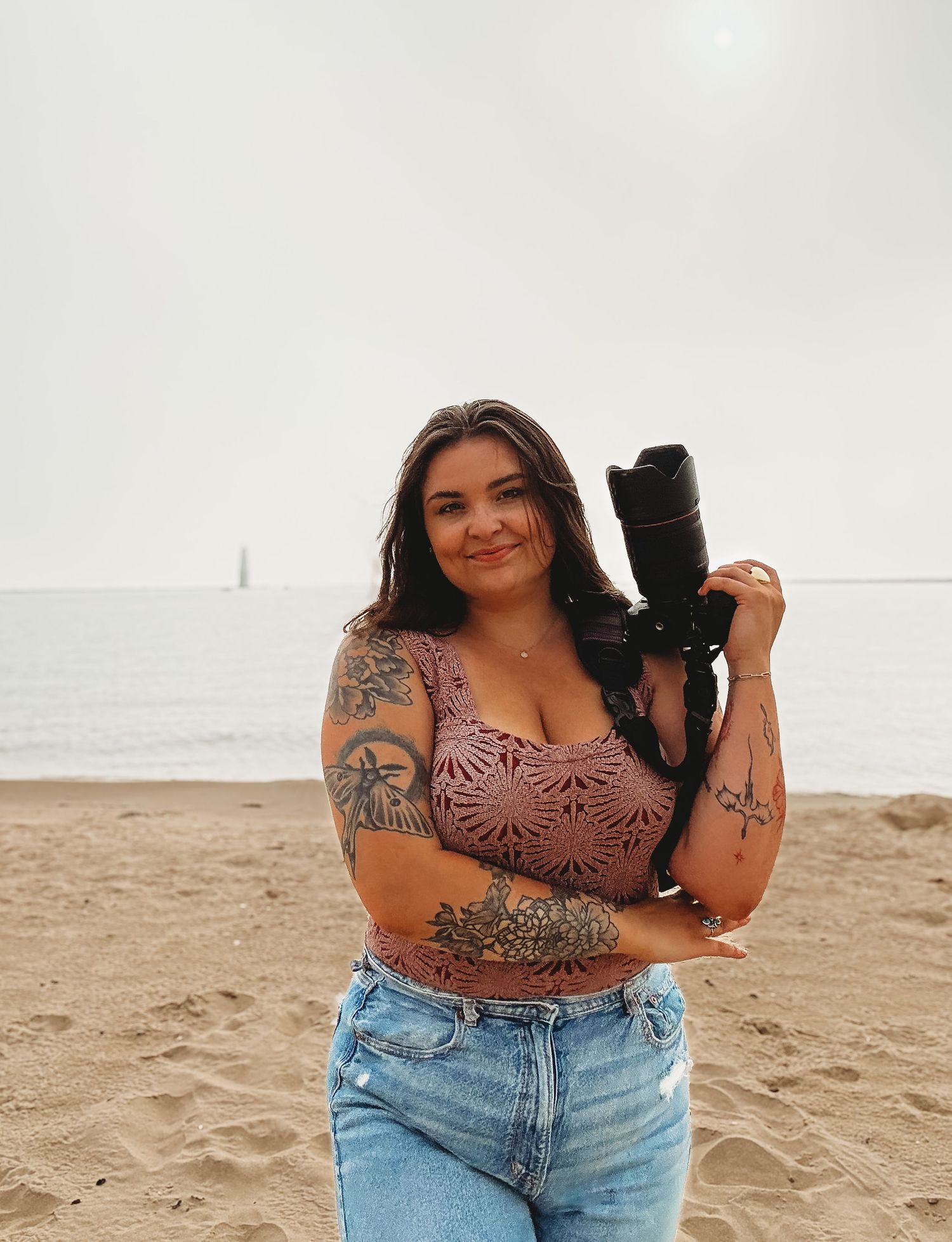 Photographer with tattoos and camera poses on sandy beach in crop top and jeans.