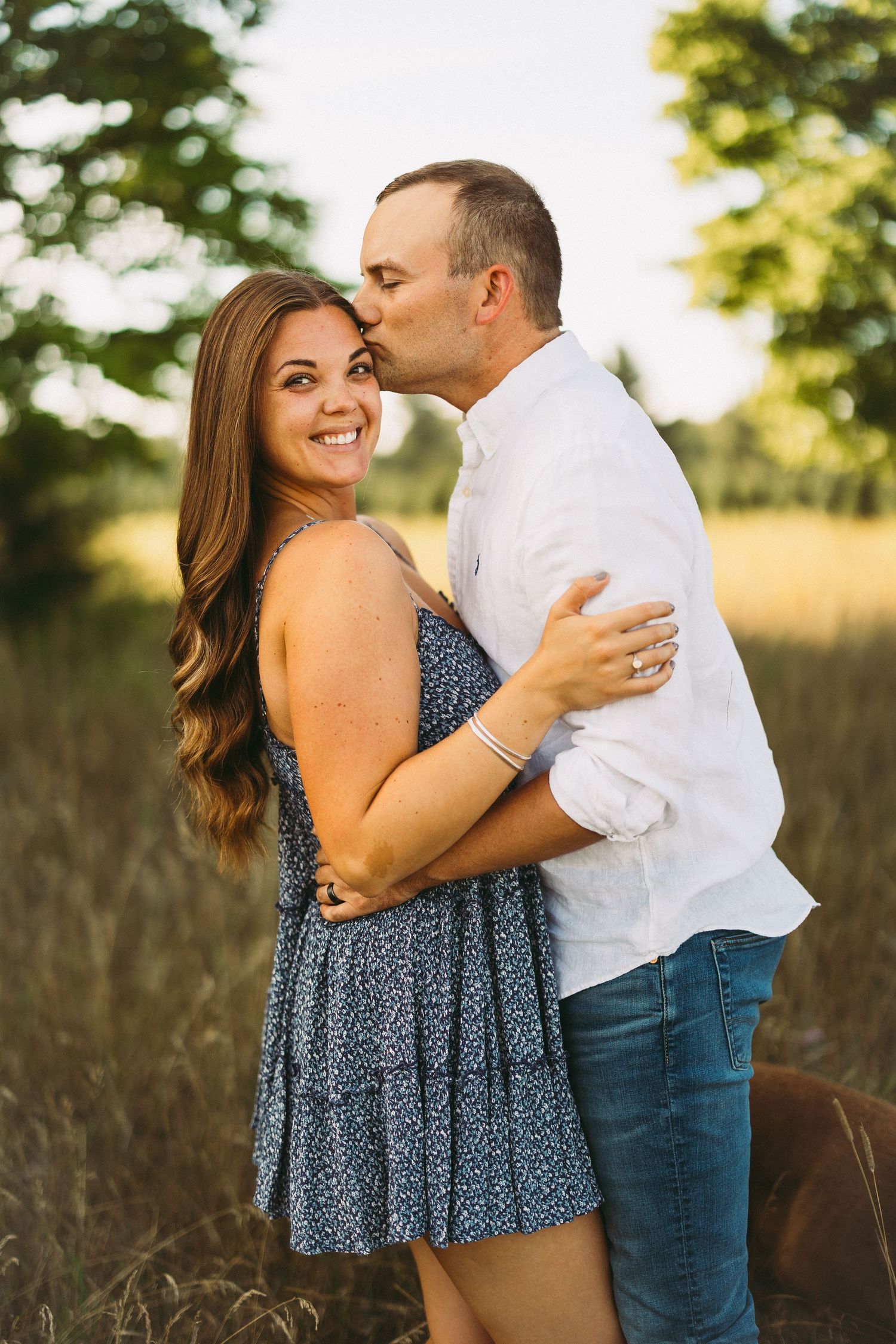 Couple in casual attire sharing intimate moments in a sunny field with trees.