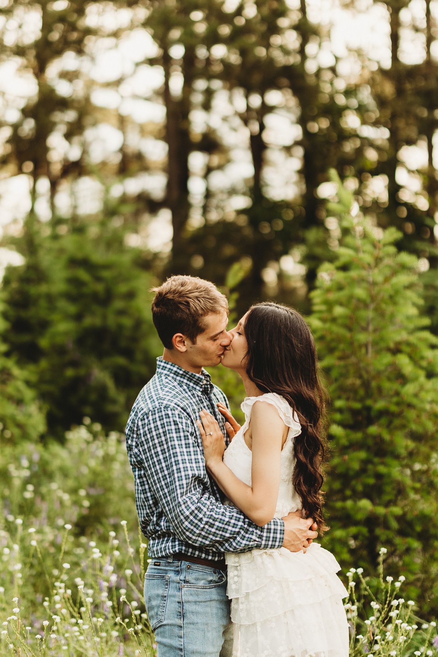 A couple shares a romantic kiss in a wildflower meadow surrounded by evergreen trees at golden hour.