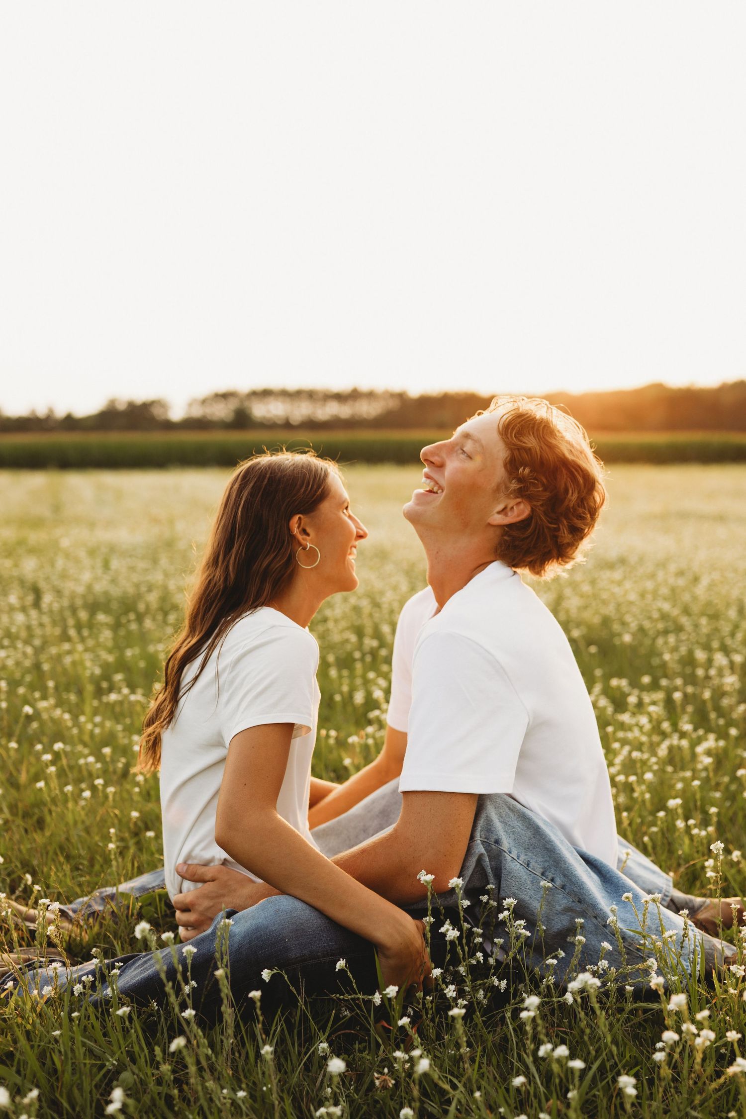 Couple sitting in a sunny flower field at golden hour, laughing and embracing in matching white t-shirts.