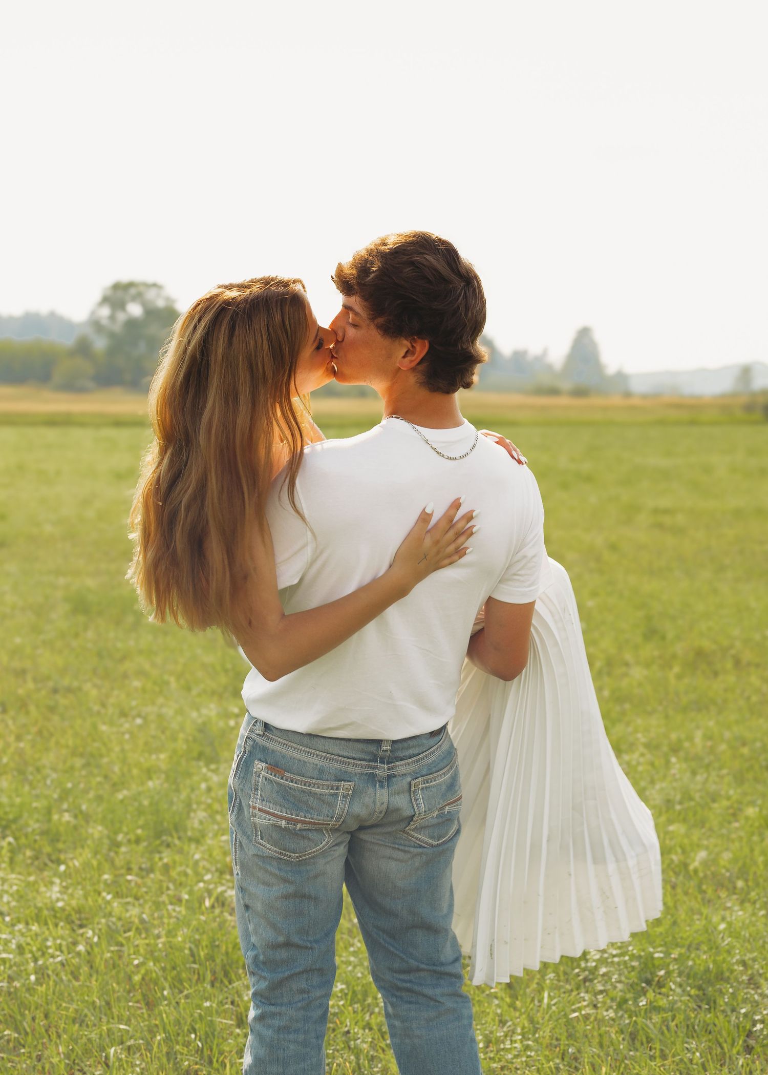 A couple sharing a romantic kiss in a sunny green field, both wearing white tops and blue jeans.