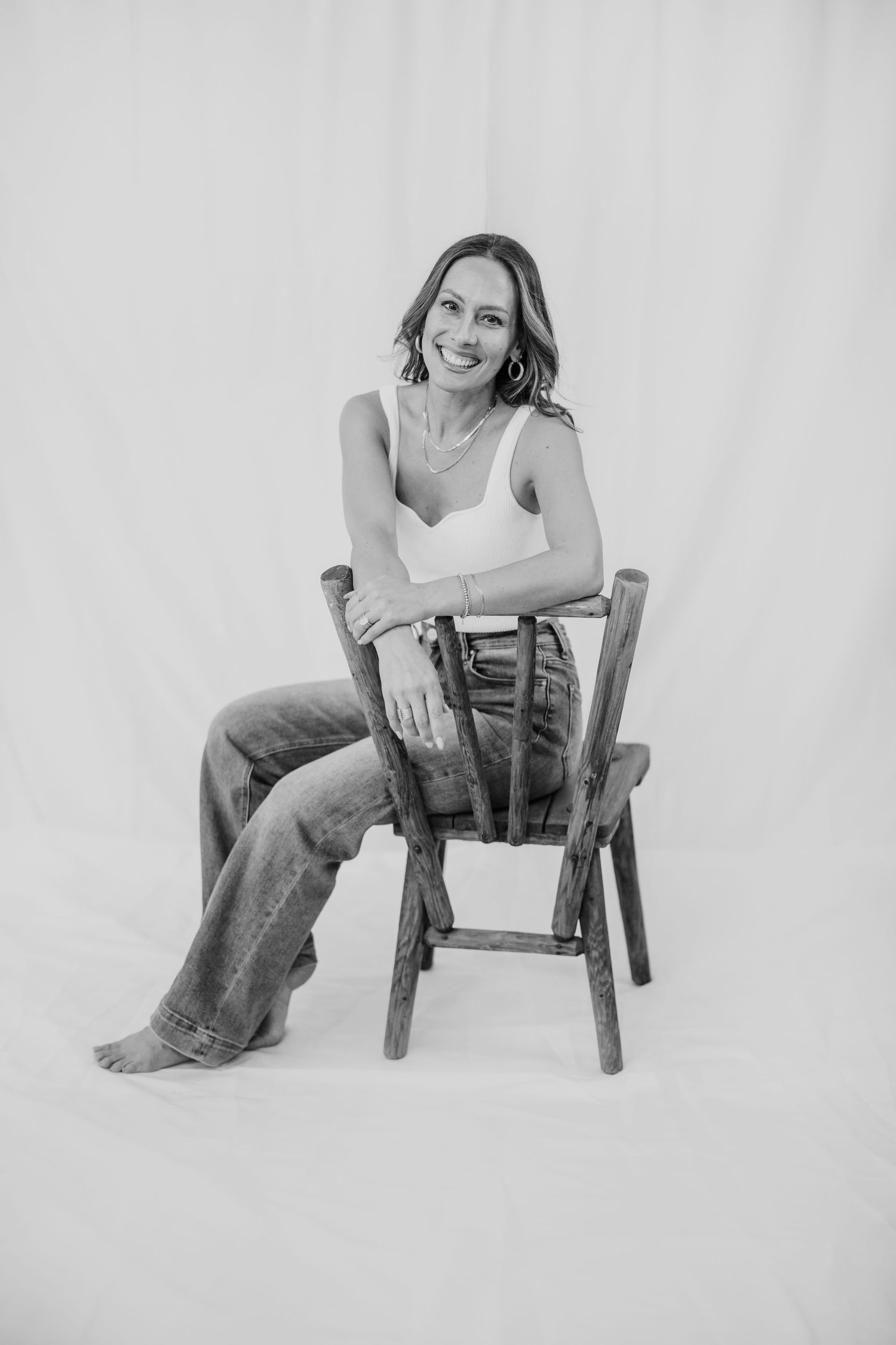 Woman smiling in white top and jeans, seated on rustic wooden chair against white backdrop, black and white photo.