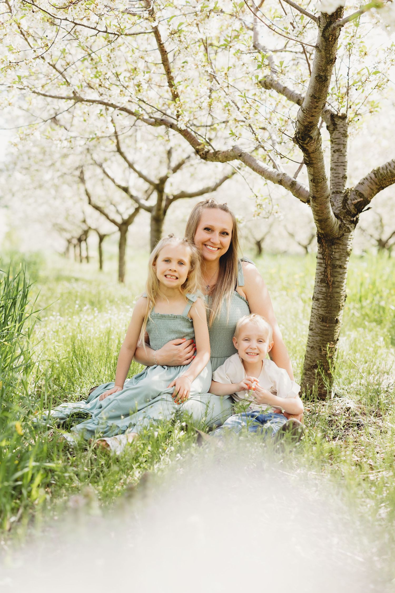 A mother and two young children sitting together under blooming spring trees in a lush green orchard.