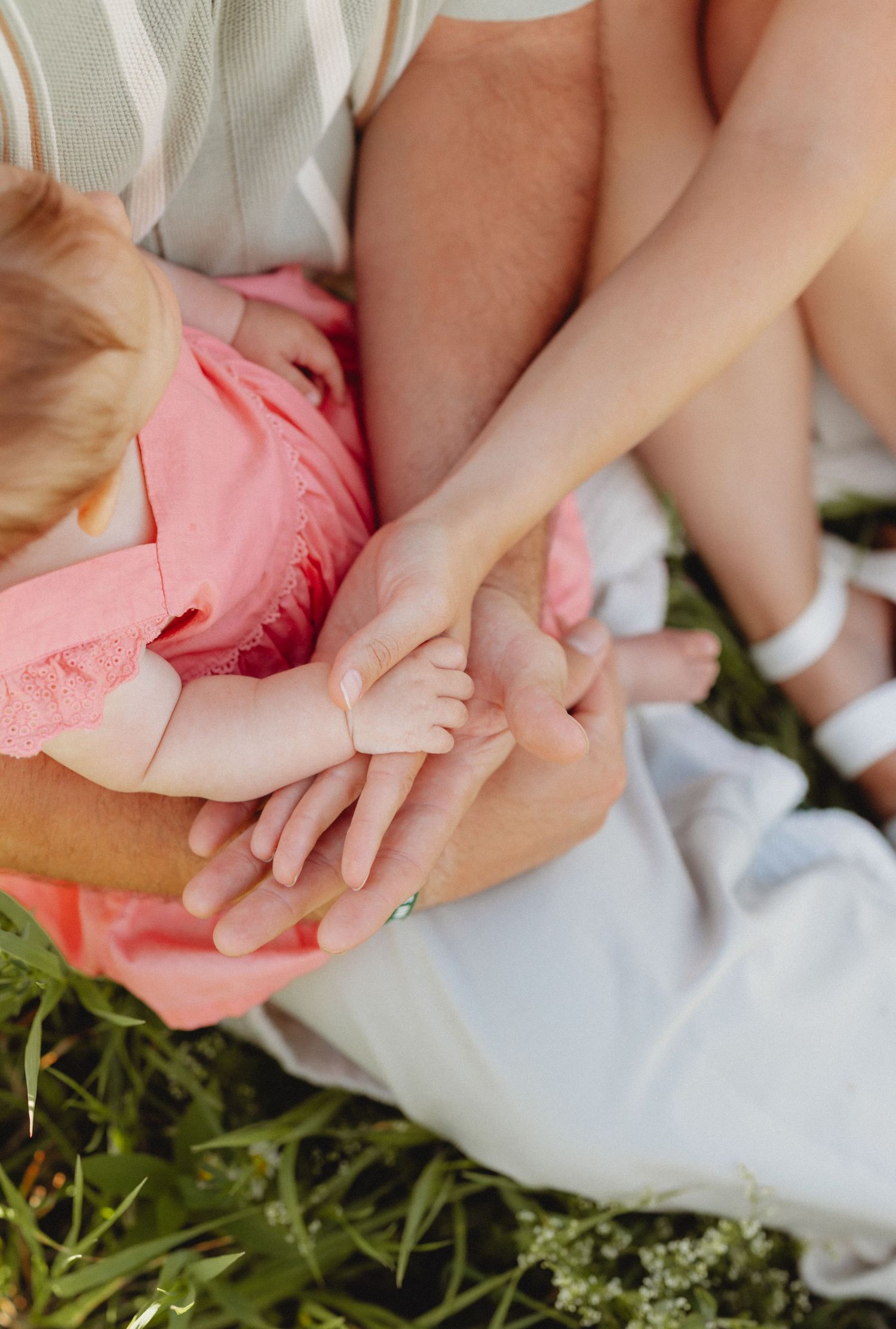 Family hands stacked together outdoors on grass, showing parents and baby bonding moment in soft natural light.
