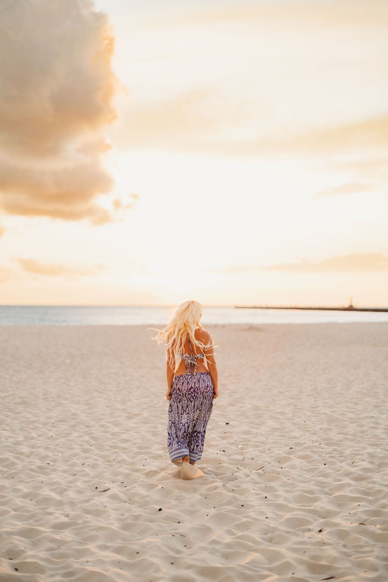 Woman with long blonde hair walks barefoot toward the ocean on a sandy beach at golden sunset.
