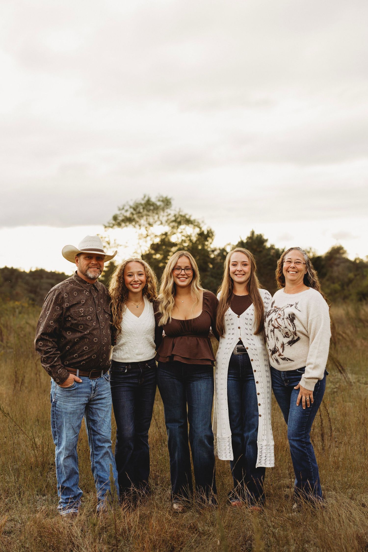 Family of five posing together in a tall grass field at sunset, wearing jeans and casual fall outfits, with a man in a cowboy hat.