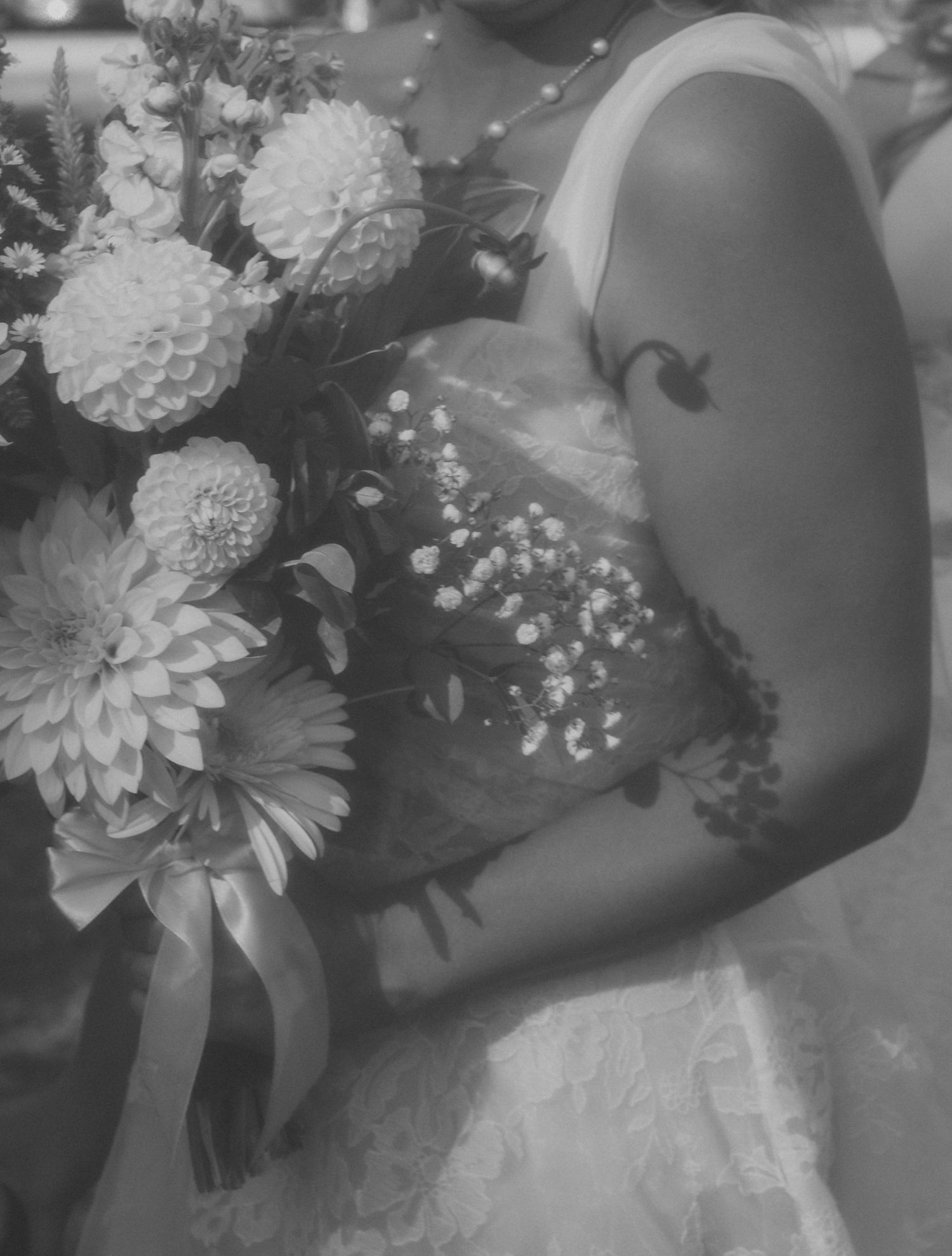 Black and white photo of a bride in a lace wedding dress holding a dahlia bouquet with a ribbon, showing her floral arm tattoo.