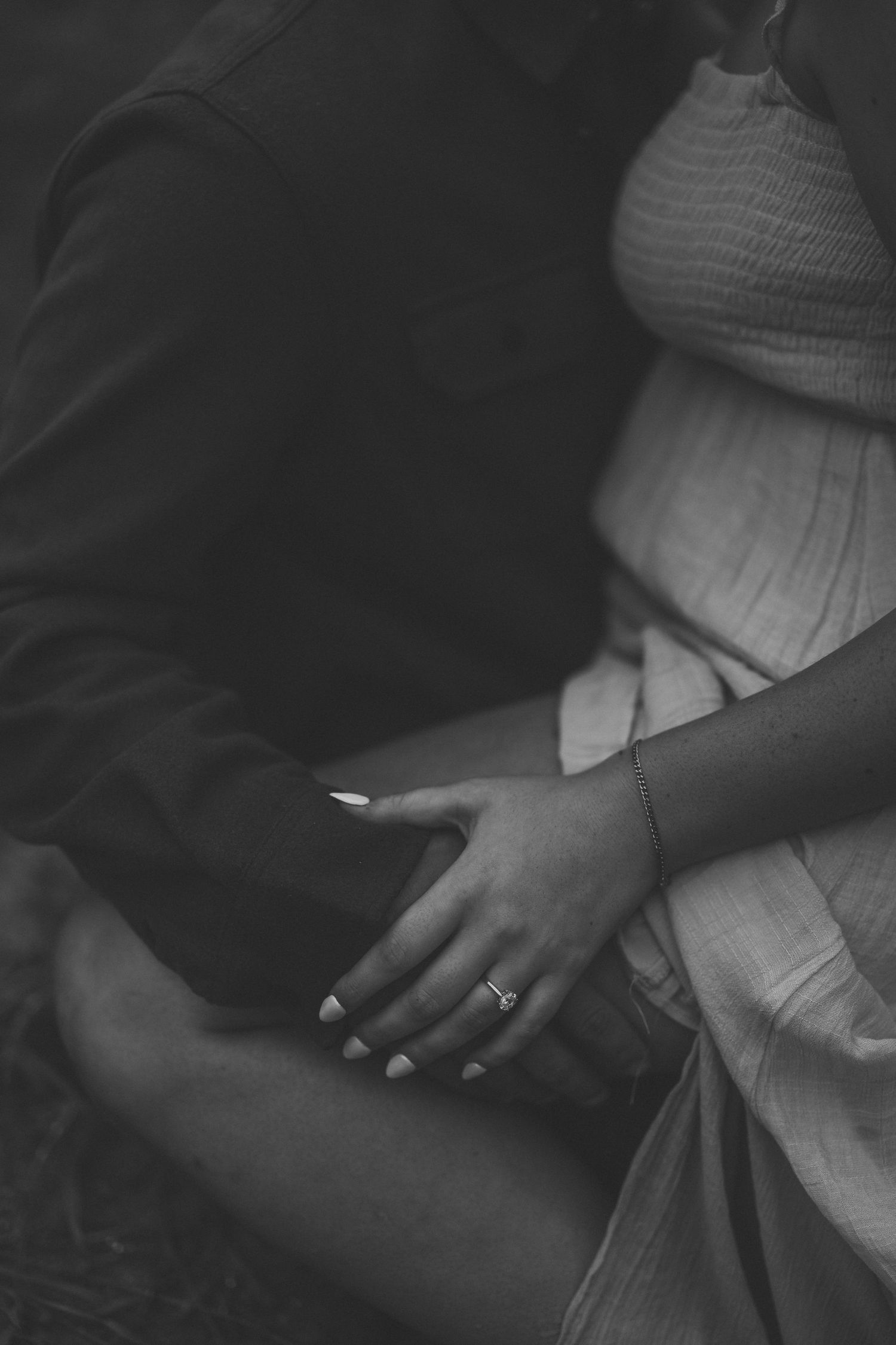 Black and white close-up of a pregnant woman in a smocked dress resting her ringed hand on a partner's arm during a maternity photo session.