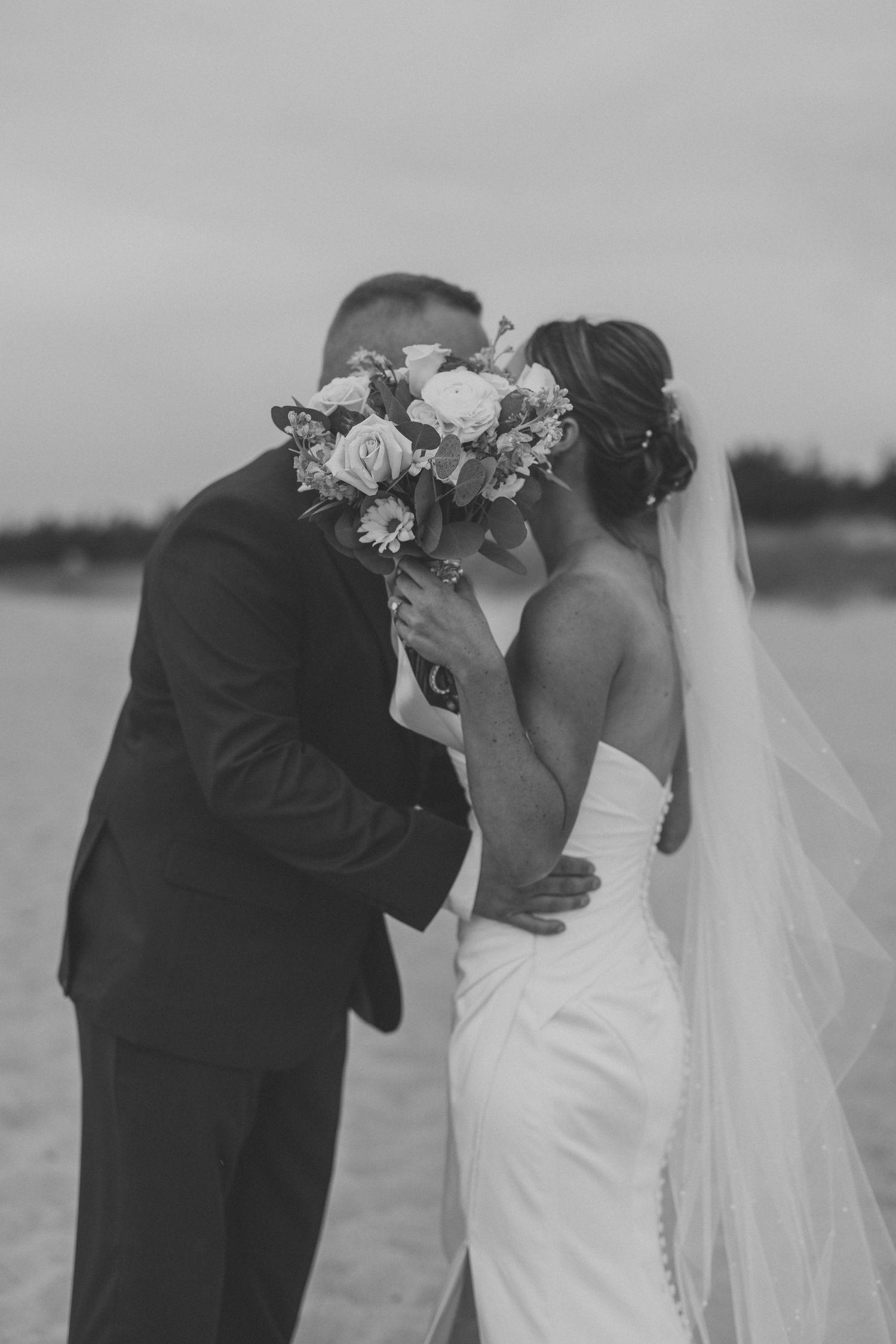 Black and white photo of bride and groom kissing behind a floral bouquet on a beach wedding day.