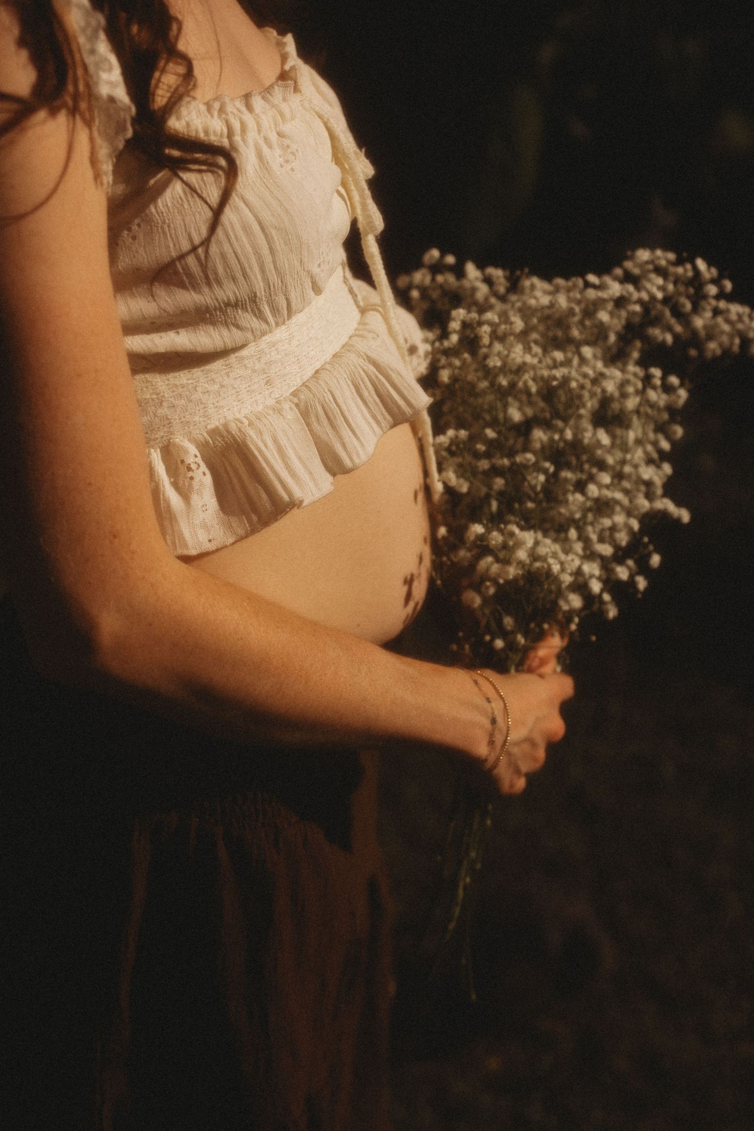Pregnant woman in white crop top holding baby's breath flowers, showcasing baby bump in warm golden light.