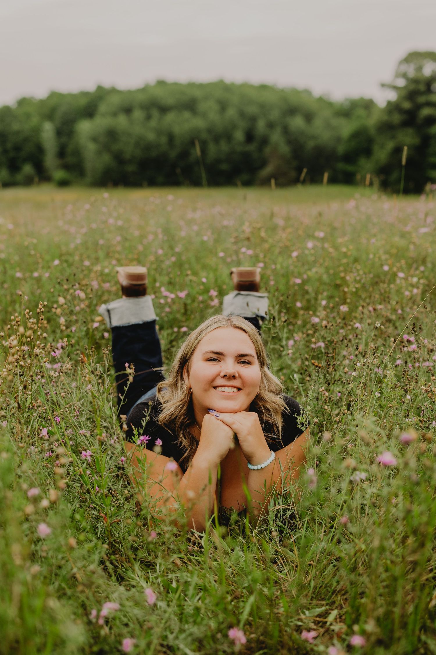 Young blonde woman lying in a wildflower meadow, smiling with chin resting on hands, green trees in background.
