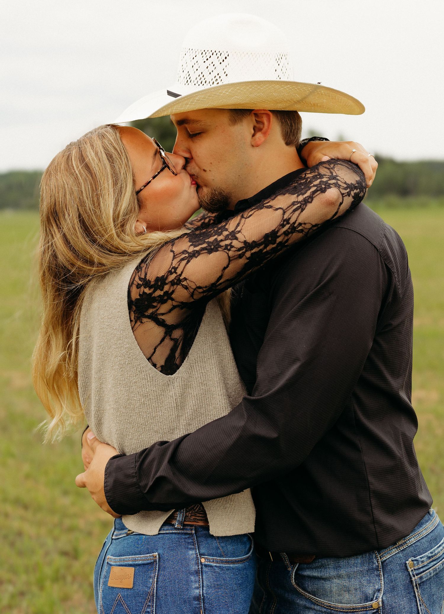 Couple sharing a romantic kiss in a green field, woman in lace sleeves embracing man in cowboy hat.