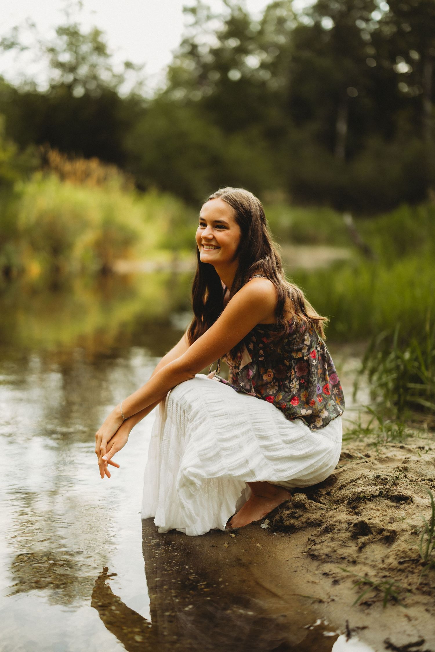 Smiling young woman with long brown hair crouching by a calm creek in a floral top and white skirt, surrounded by green trees.