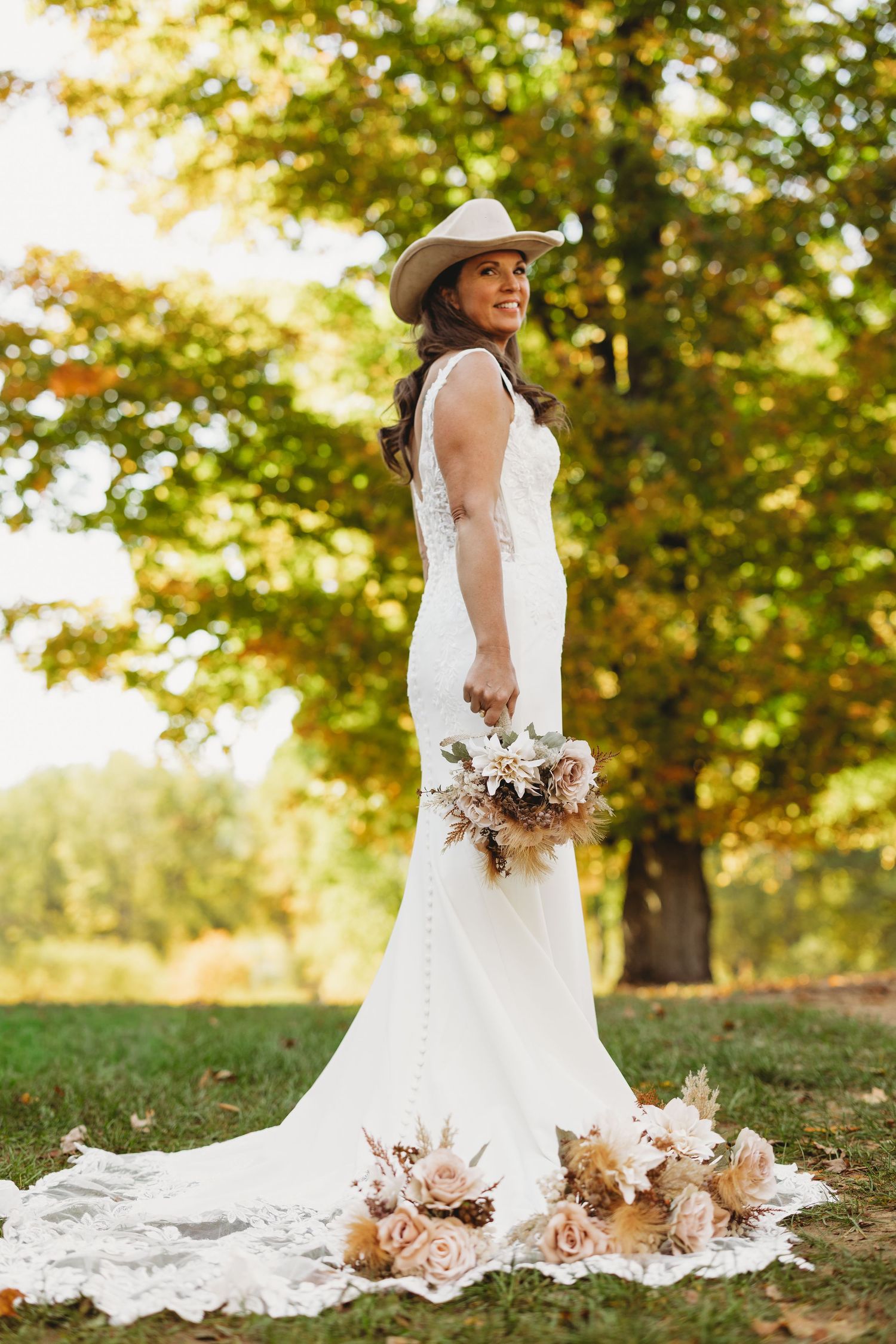 Smiling bride in a lace fitted wedding gown and tan cowboy hat holds a dried floral bouquet on grass beneath autumn trees.