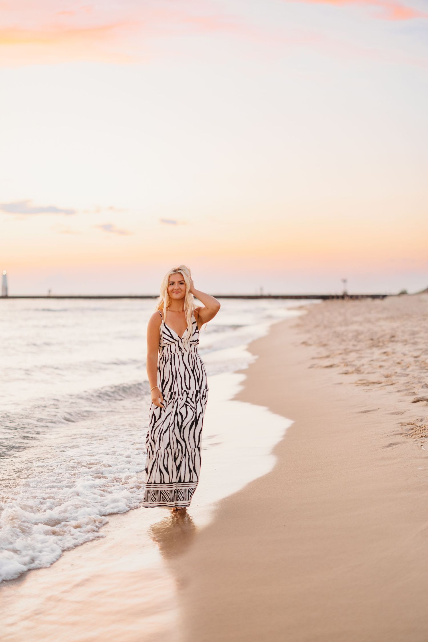 Woman in a black and white zebra print maxi dress walking along the shoreline of a sandy beach at sunset with soft pink skies.
