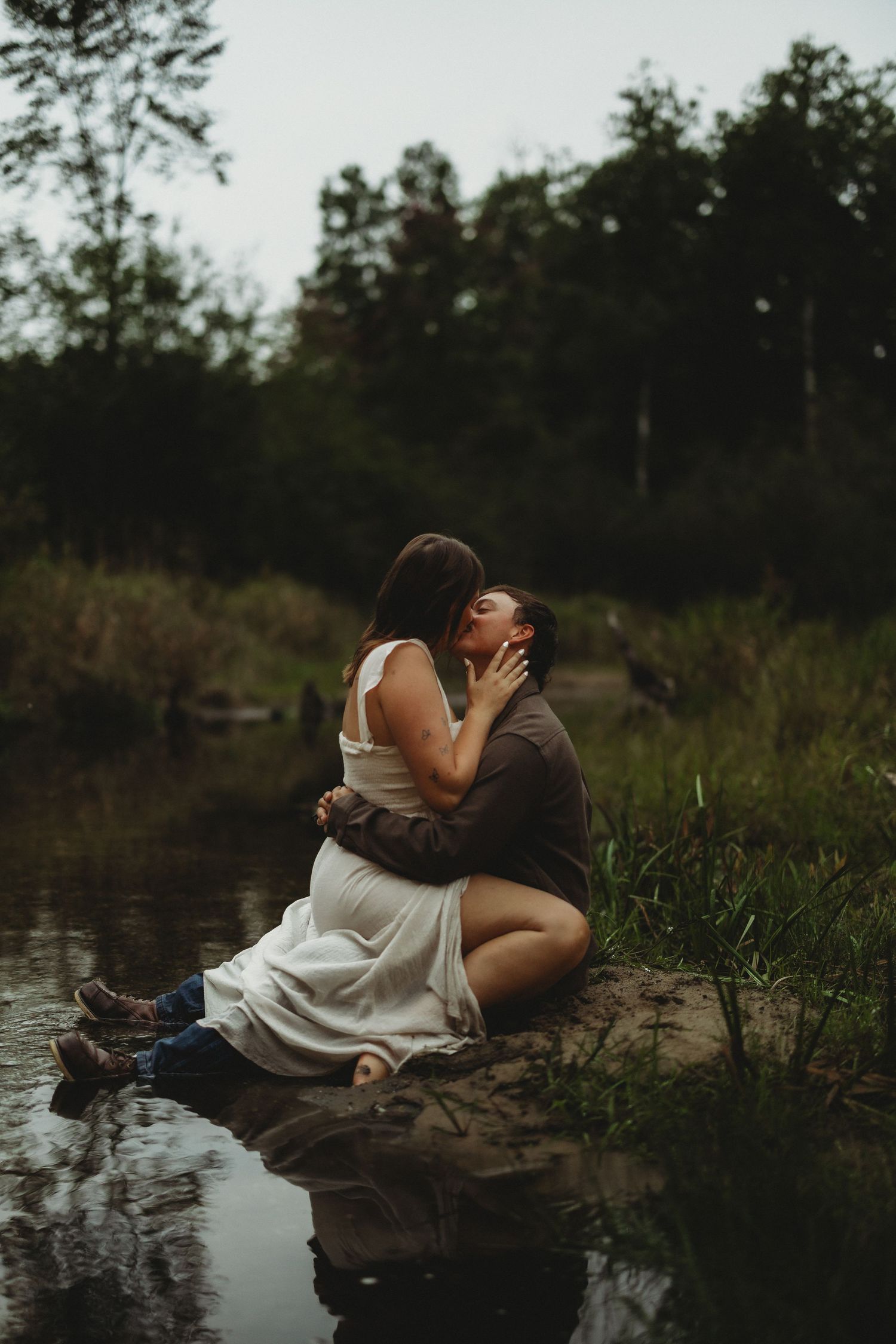 Couple in a white dress and brown shirt embrace and kiss on a rock beside a calm forest pond at dusk.