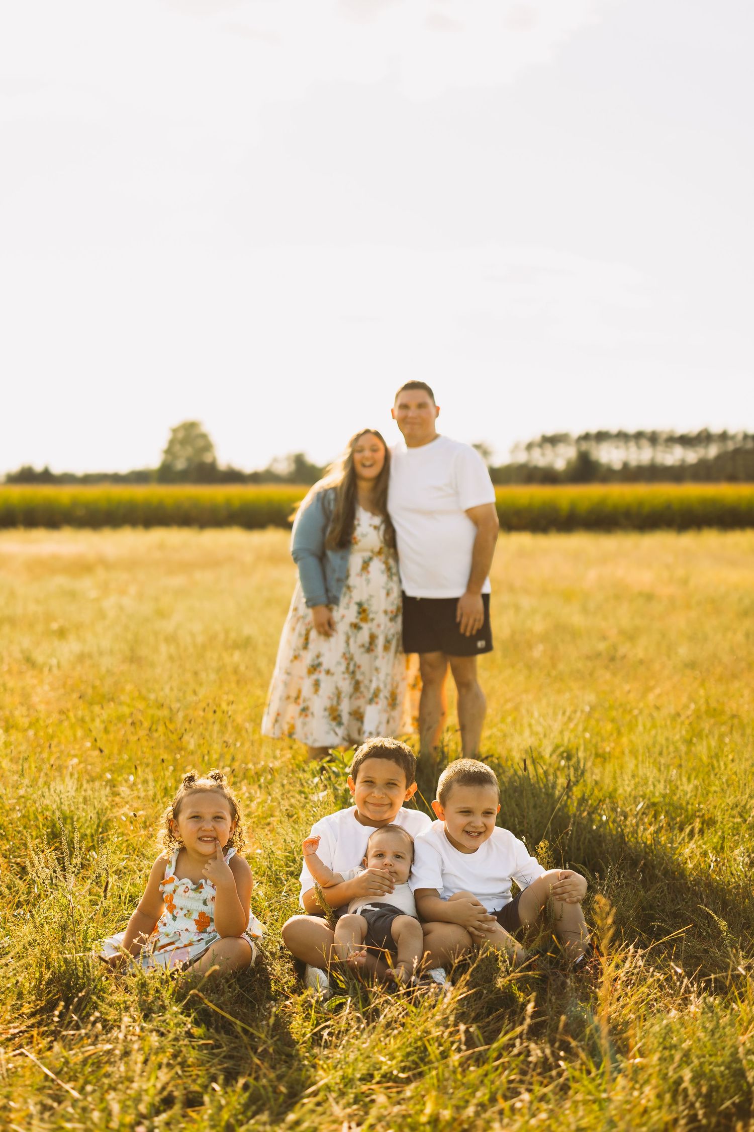 Smiling family of six posing together in a golden grassy field at sunset, with parents standing behind four children seated in the grass.