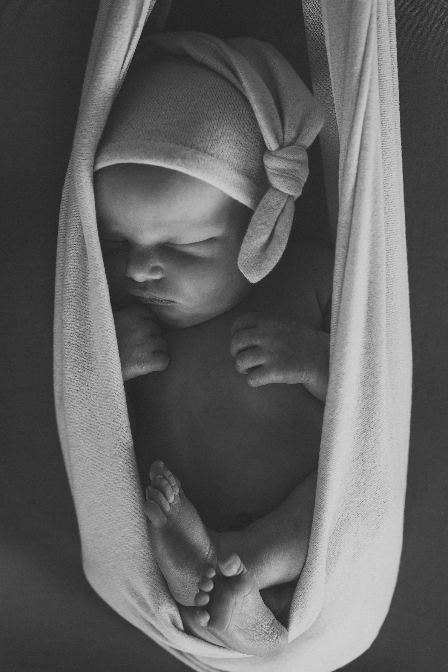 Black and white photo of a sleeping newborn baby wrapped in soft fabric, wearing a knotted hat.