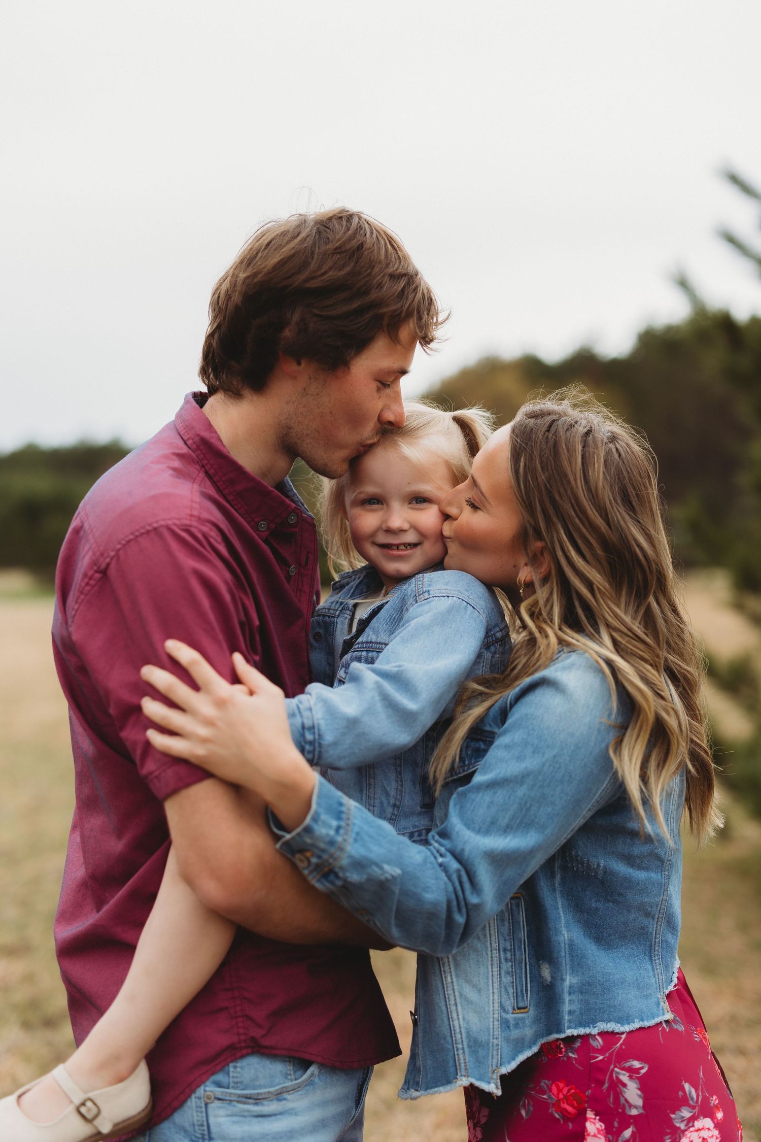 A young family shares a tender moment outdoors, parents kissing their smiling toddler on the cheeks in a field.