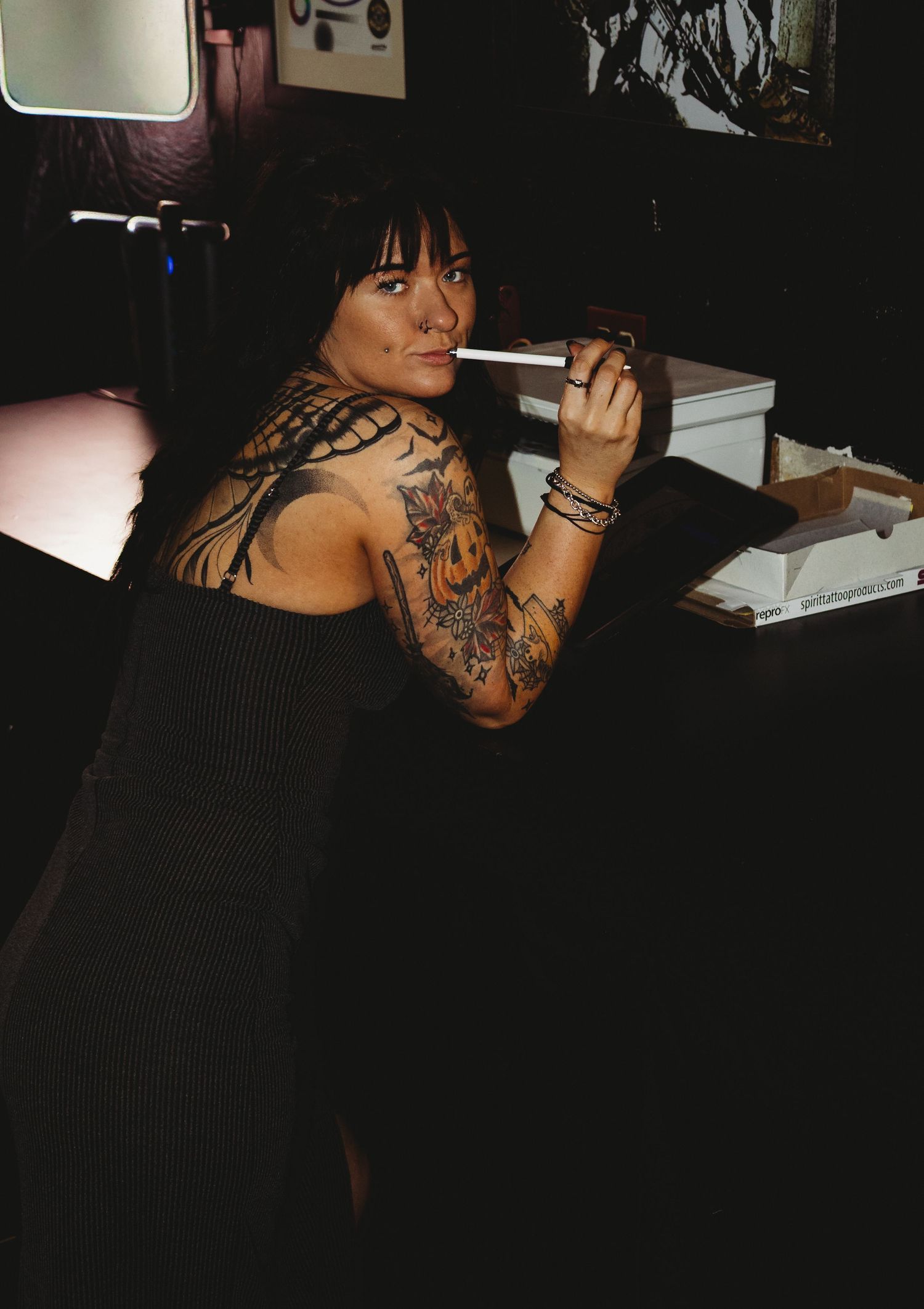 Tattooed woman in black dress smoking a cigarette at a dimly lit desk, showcasing extensive arm and neck tattoos.