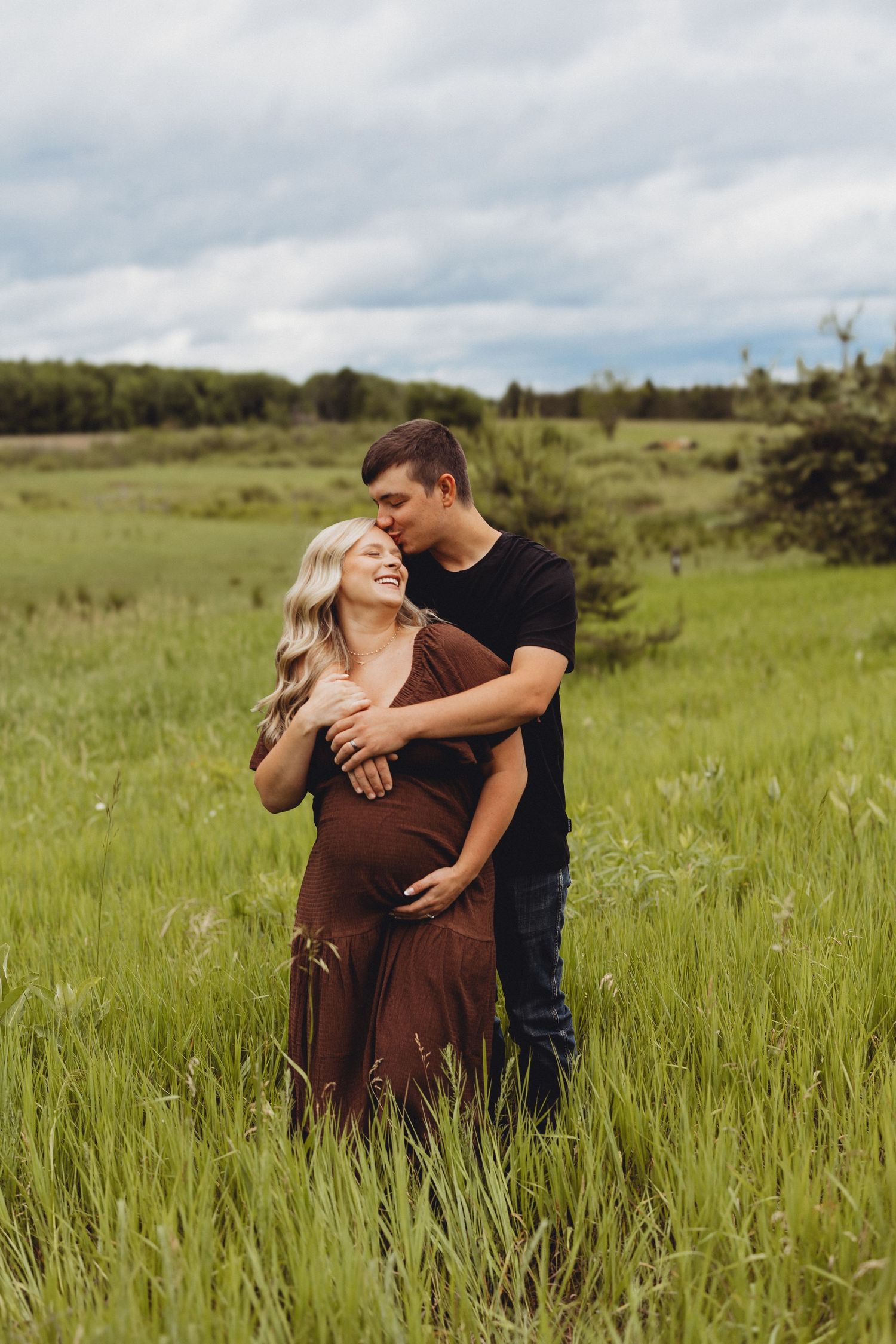 Couple sharing a tender moment in a lush green field, pregnant woman in brown dress smiling as man kisses her cheek.