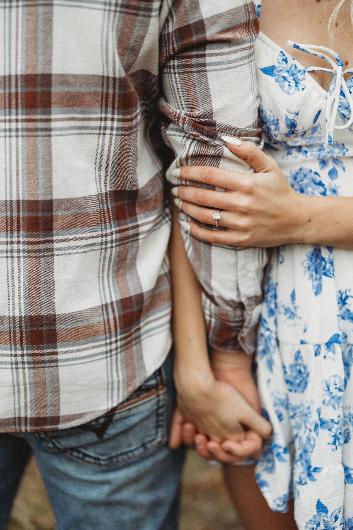 Close-up of couple holding hands, wearing plaid shirt and blue floral dress with engagement ring visible.