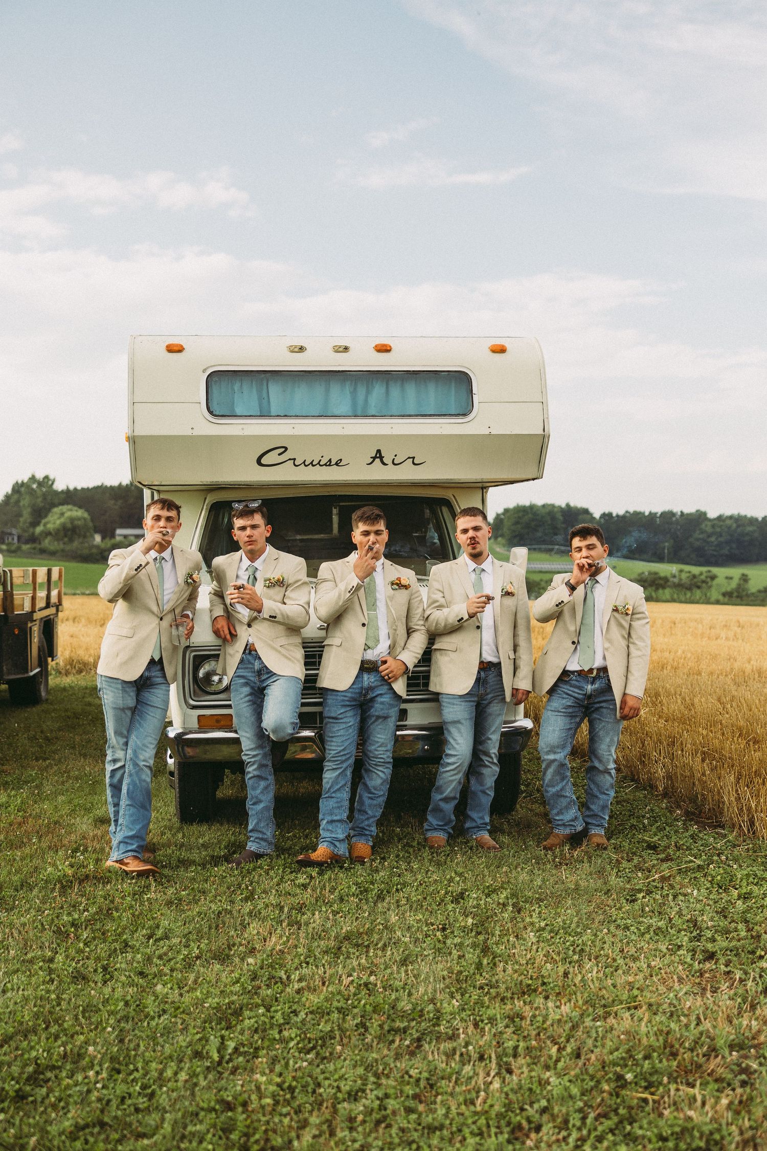 Five people in matching denim and white shirts pose playfully behind a vintage Cruise Air camper trailer in a rural field.