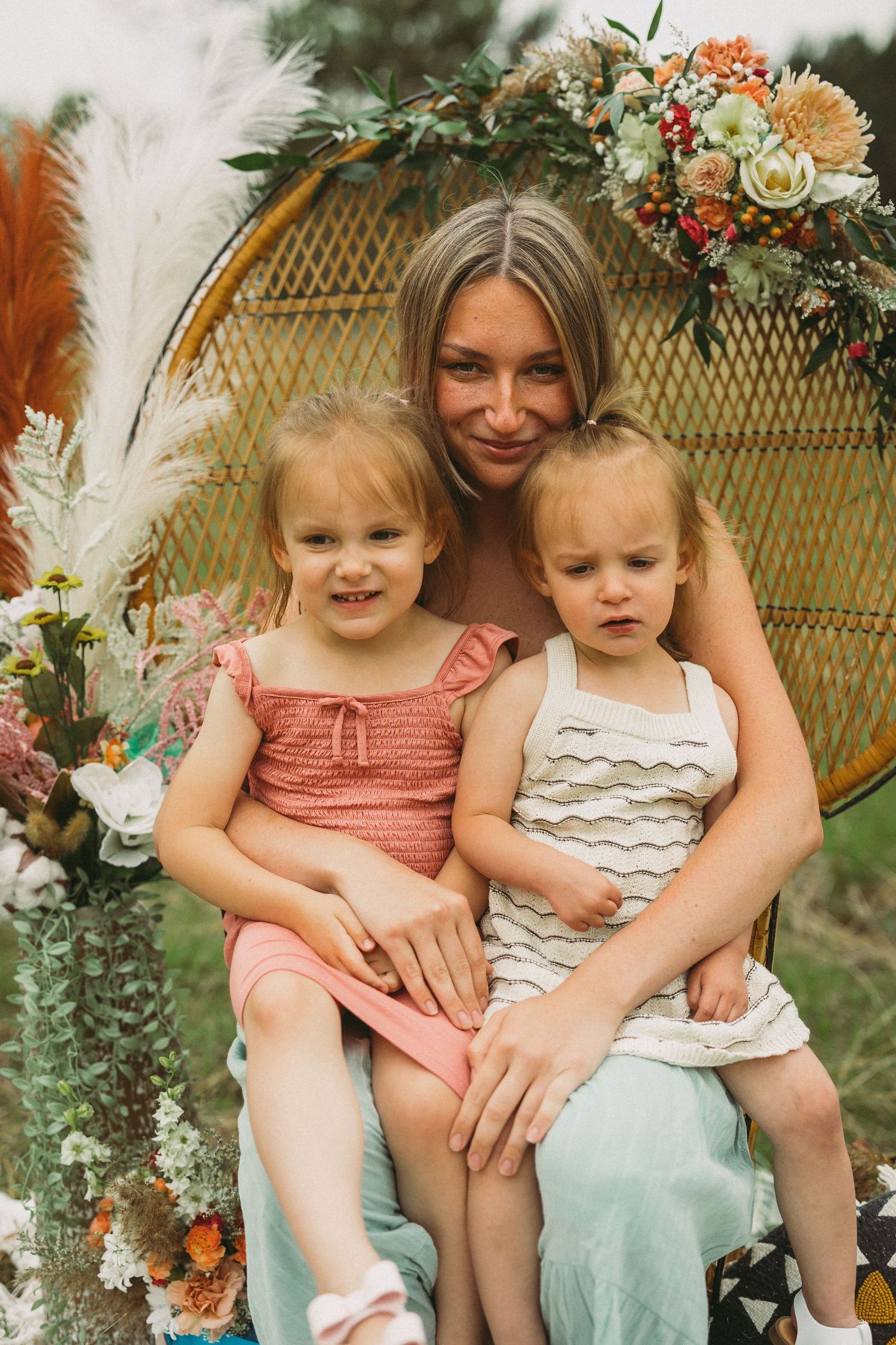 Two children sit together in a wicker peacock chair surrounded by floral decorations and eucalyptus.
