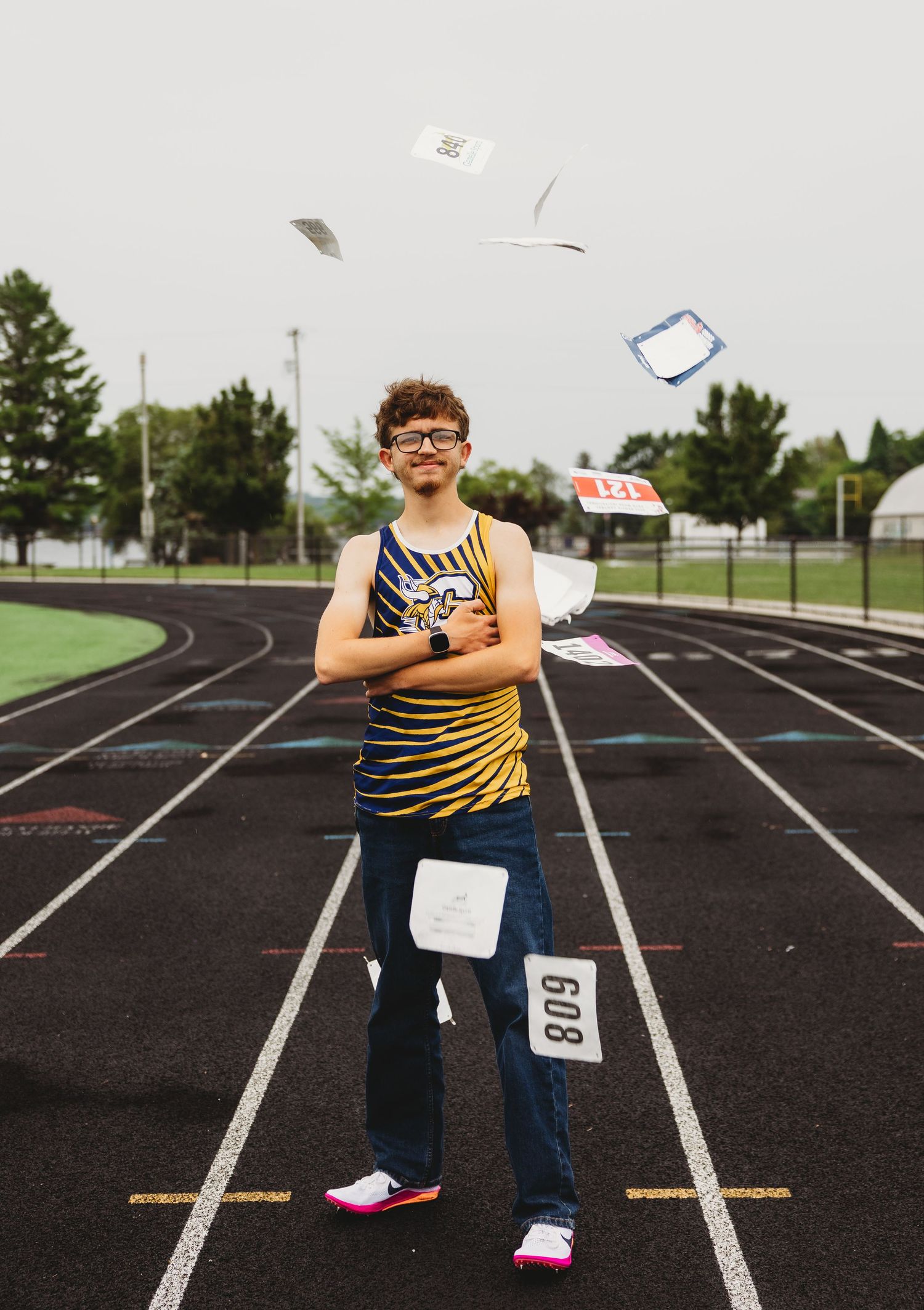 Smiling track athlete in a striped jersey and jeans stands with arms crossed on a running track as race bibs and papers fly through the air.