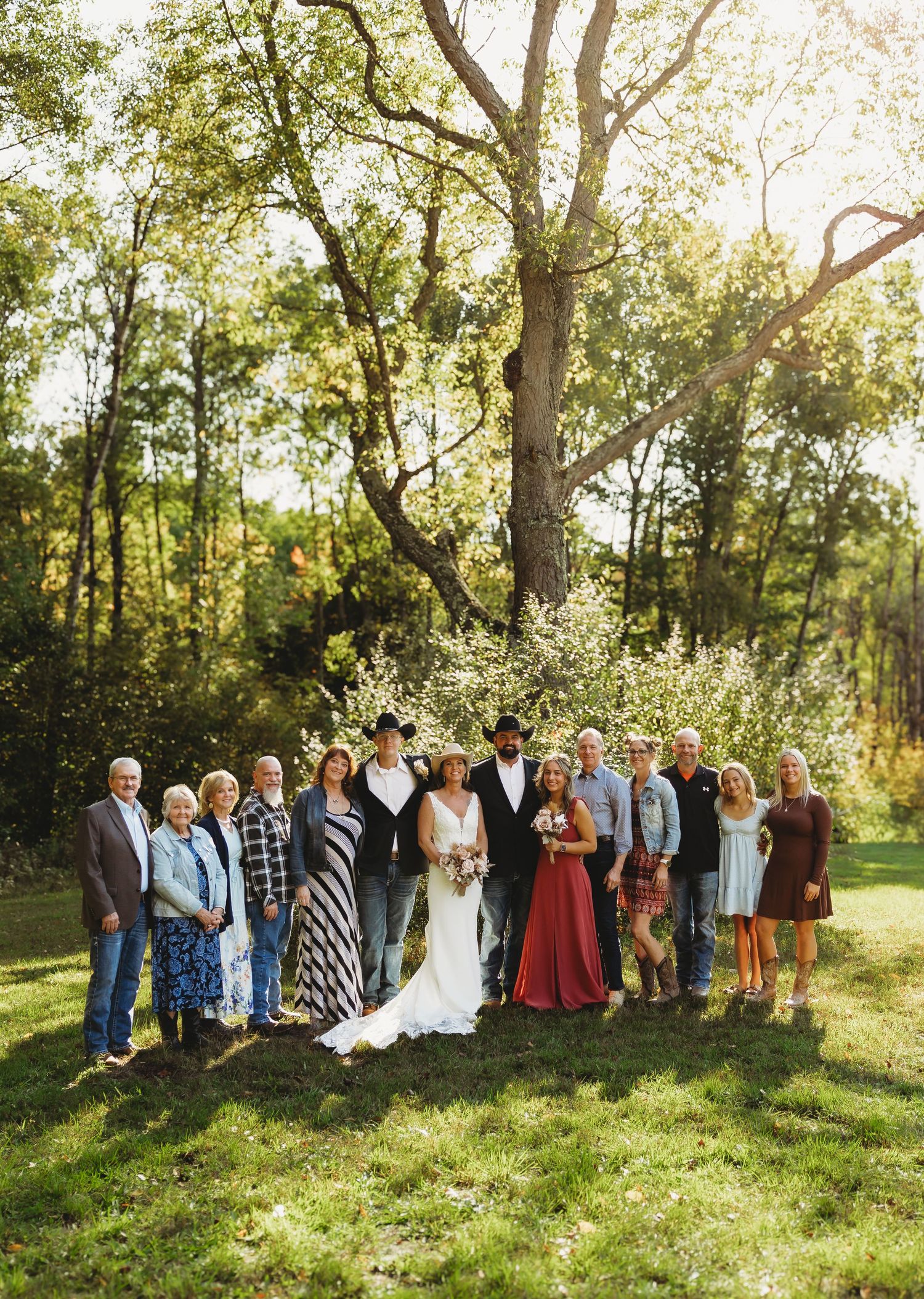 Wedding party and family pose outdoors on a sunny grassy lawn beneath tall trees, with the bride in a white gown and groom in a cowboy hat.