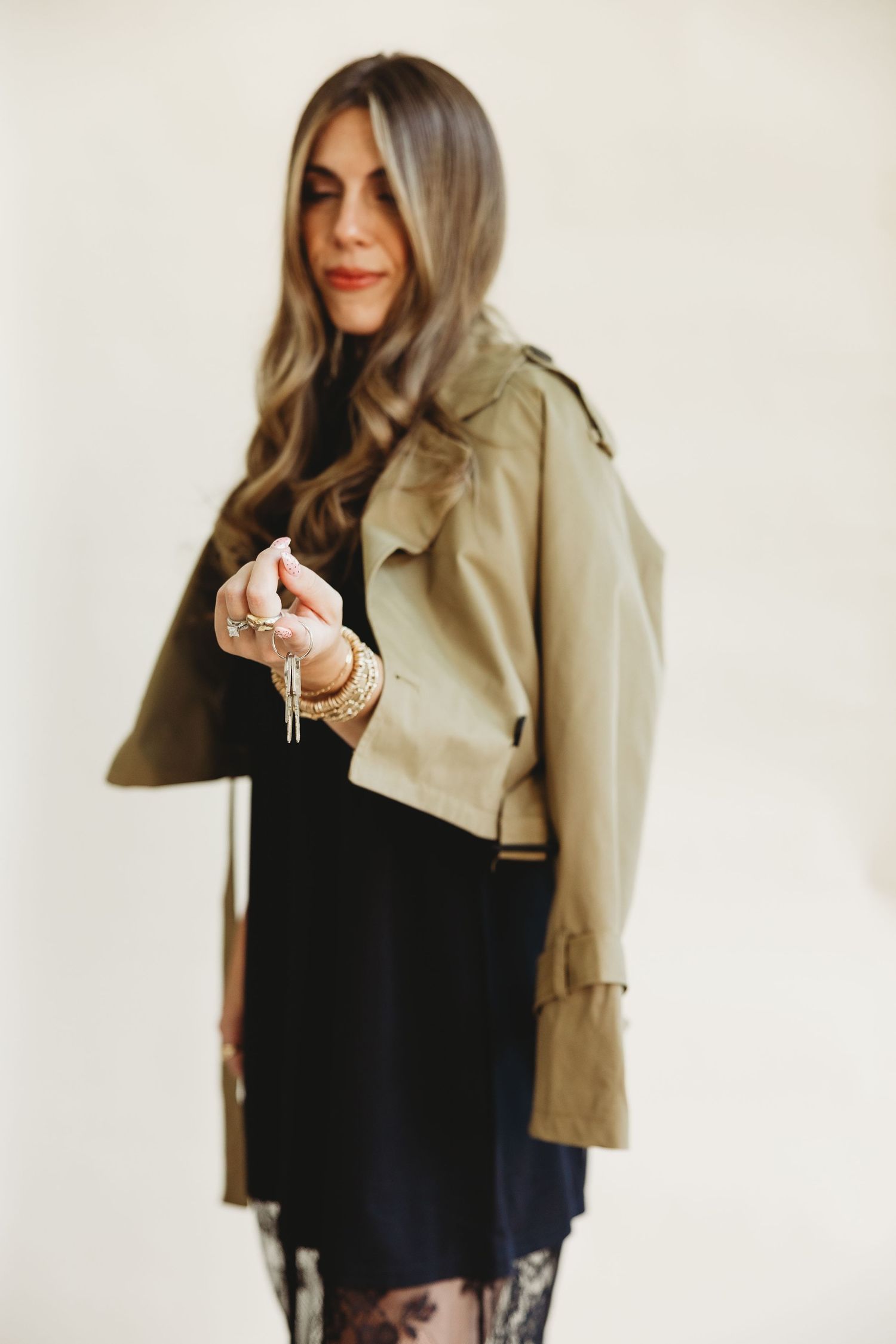 Woman with long wavy highlighted hair wearing a khaki cropped trench jacket, black dress and lace tights while holding a tassel necklace.