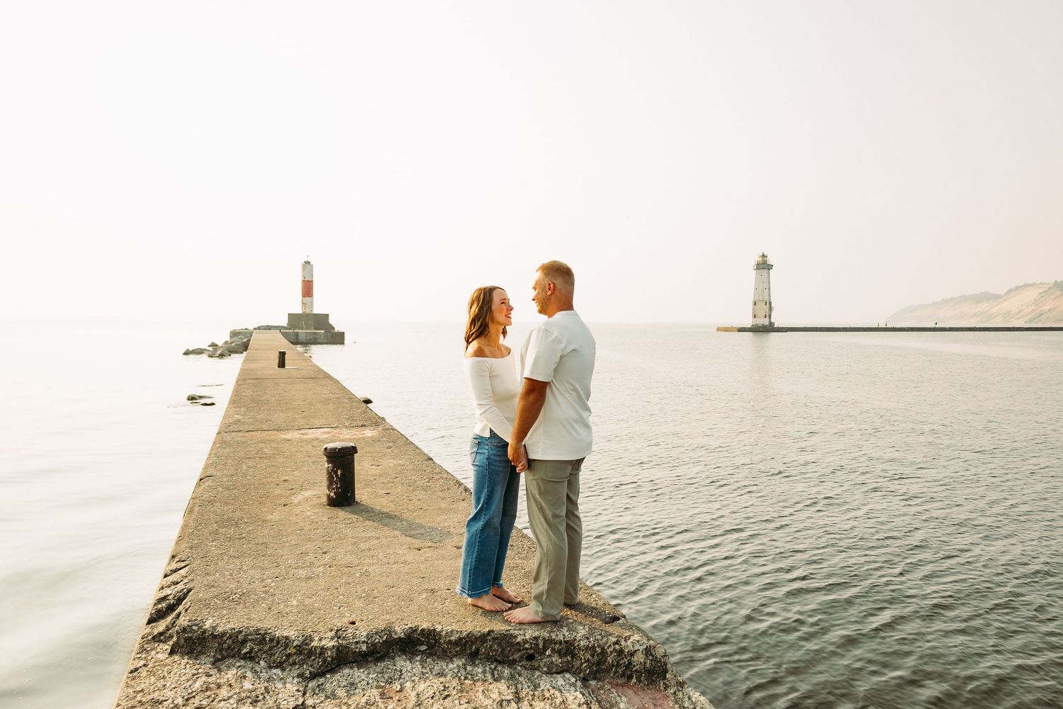 Couple holding hands on a stone pier with two lighthouses and calm sea in the background at sunset.