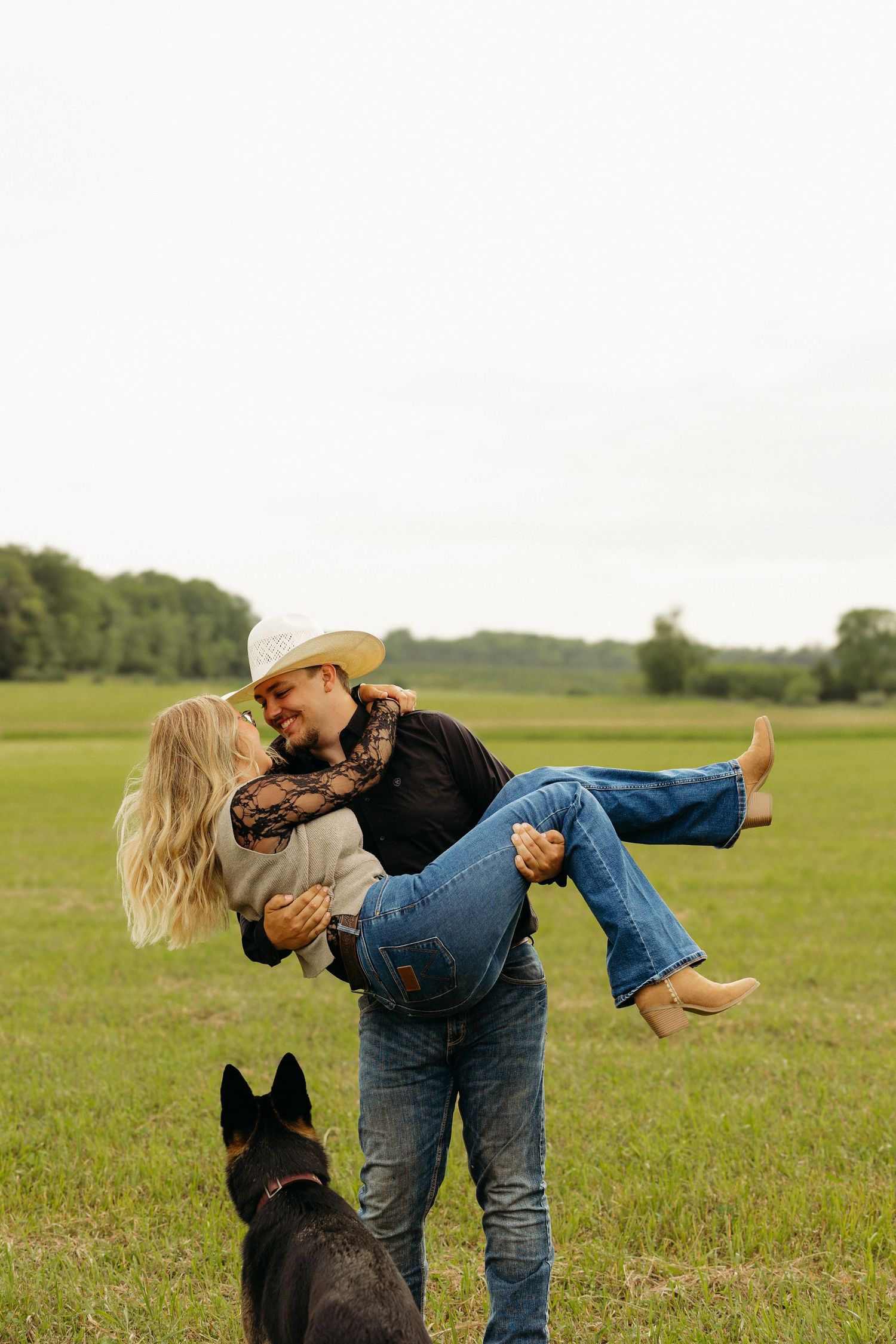 A cowboy in a white hat lifts a blonde woman in jeans and boots while a German Shepherd watches in a green field.