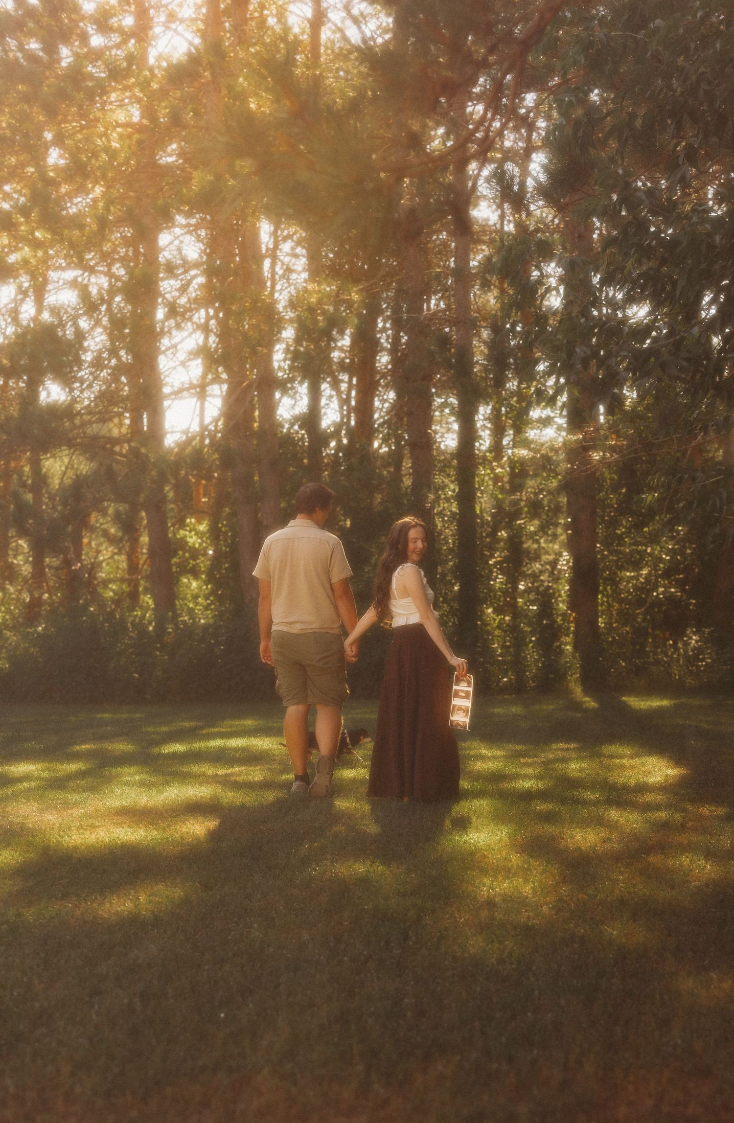 Couple holding hands walking toward sunlit forest on green grass, woman in maxi skirt carrying basket.