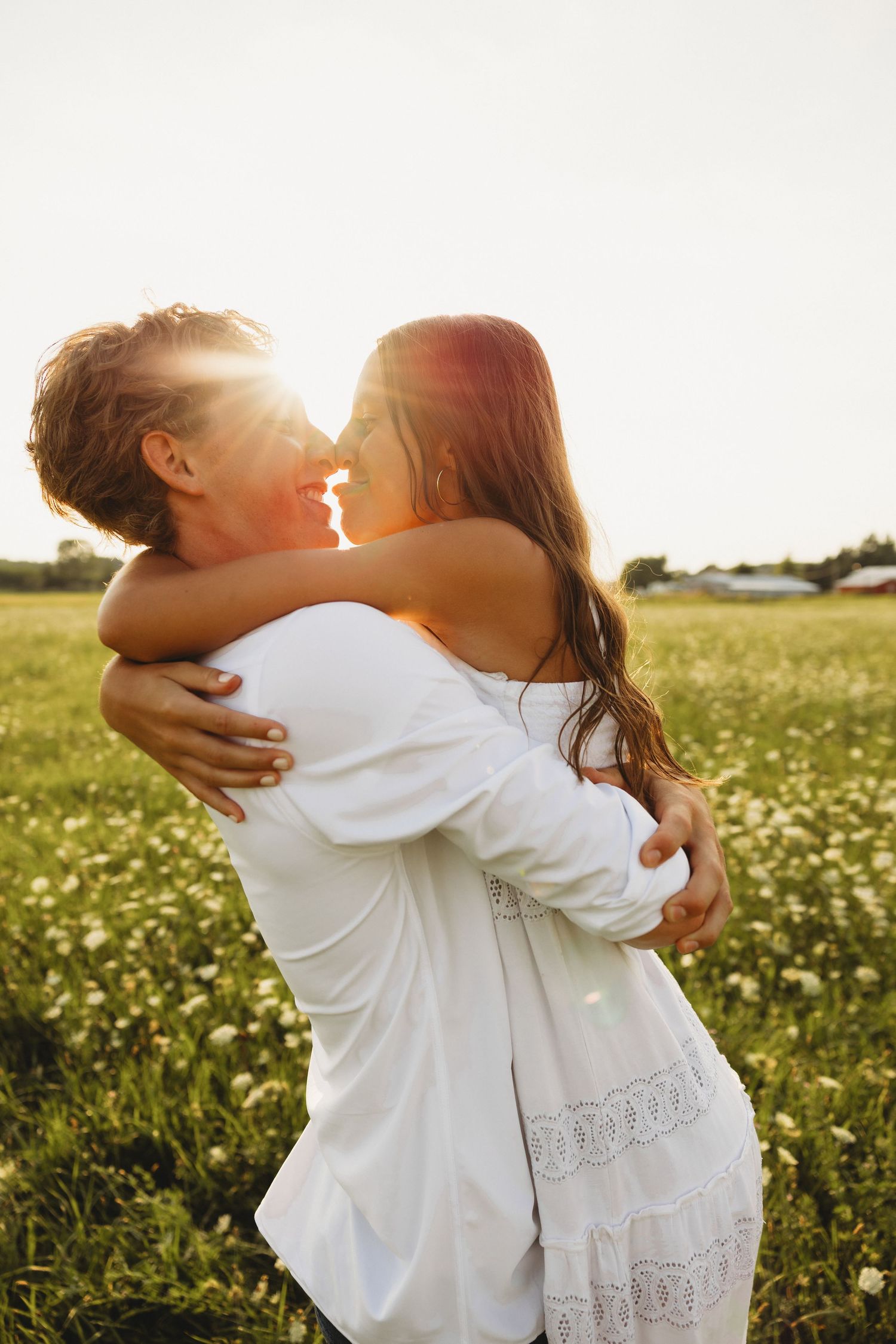 Couple embracing and kissing in a sunny flower field, backlit by golden sunset light, both wearing white.