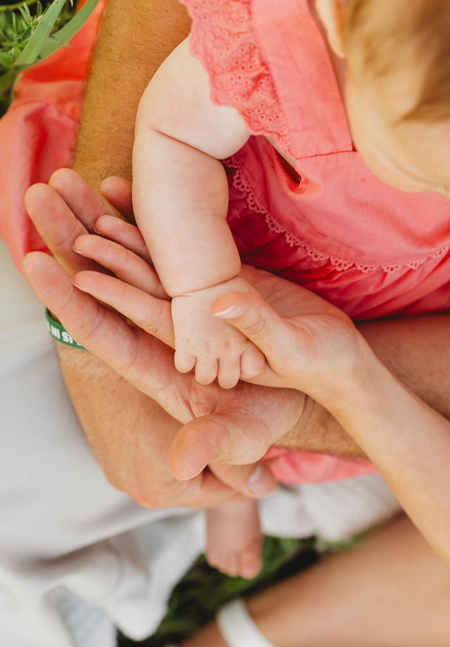 A newborn baby's tiny hand gently gripping an adult's finger, with parents in coral outfits cradling the infant.