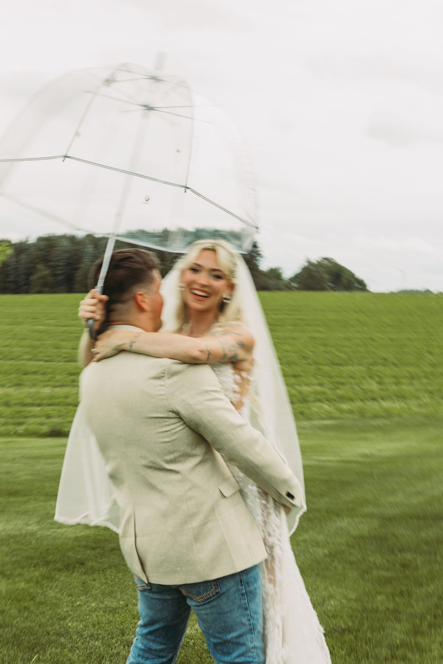 Couple shares intimate moment under clear umbrella in green field.