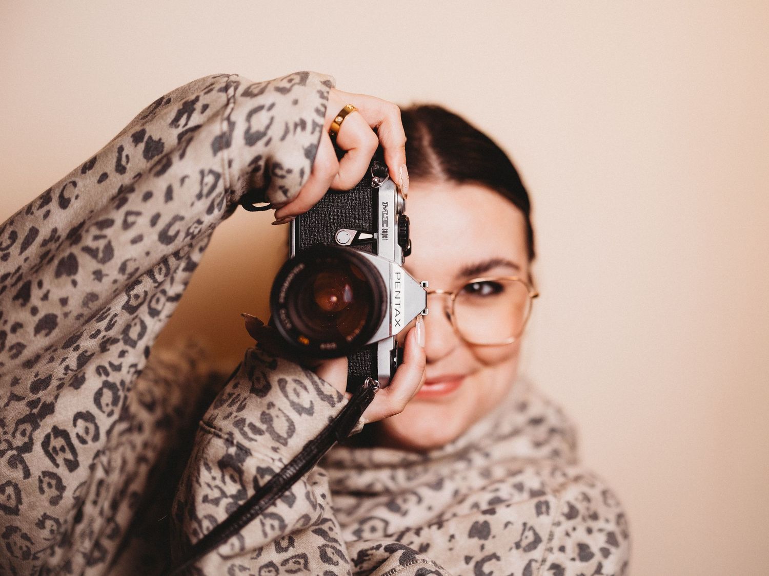 Woman in leopard print scarf holds vintage film camera up to her eye, smiling, wearing gold glasses.