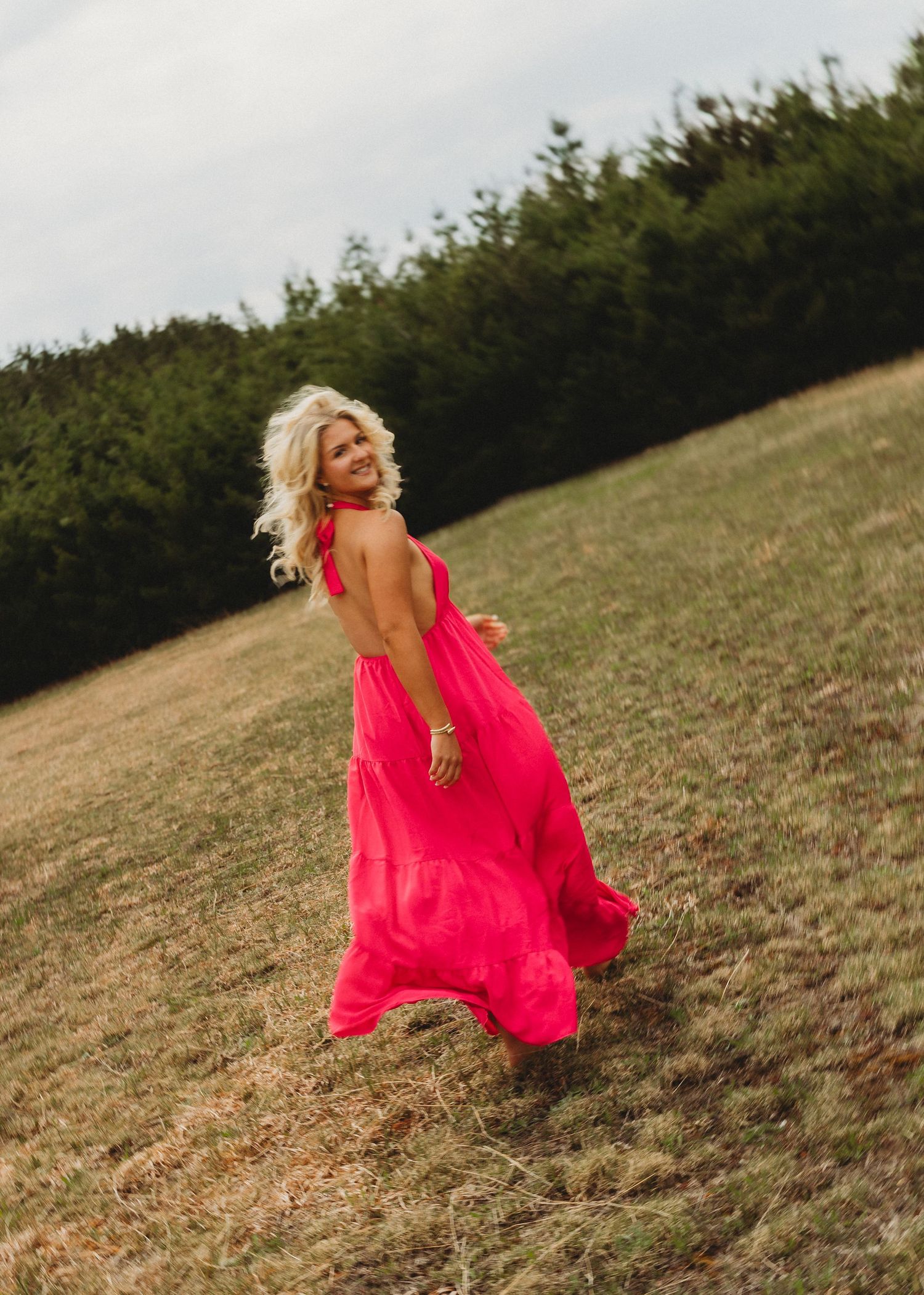 Blonde woman in flowing hot pink maxi dress twirling in a green field with trees in the background.